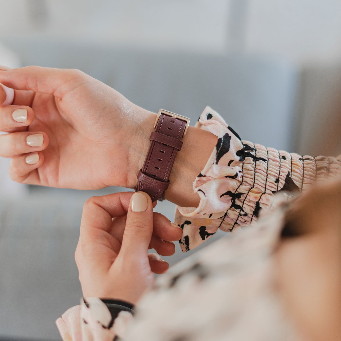 Eine Person befestigt ein kastanienbraunes Arktisband Apple Watch Lederarmband "Premium" an ihrem Handgelenk, während sie eine geblümte langärmelige Bluse trägt.