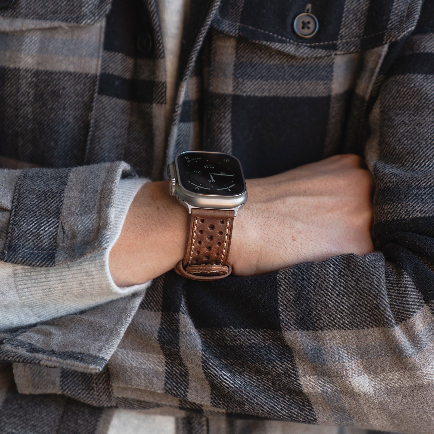 Person im karierten Hemd mit verschränkten Armen, die das arktisband Apple Watch Lederarmband "Toronto" von arktisband mit einem braunen perforierten Lederband im Vintage-Stil präsentiert.