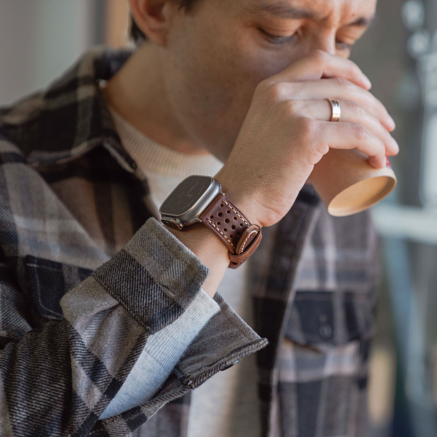 Ein Mann im karierten Hemd nippt an einem Pappbecher, trägt das arktisband Apple Watch Lederarmband "Toronto" und einen Ehering an der linken Hand.