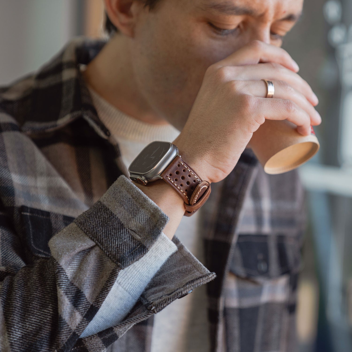 Ein Mann im karierten Hemd nippt an einem Pappbecher, trägt das arktisband Apple Watch Lederarmband "Toronto" und einen Ehering an der linken Hand.