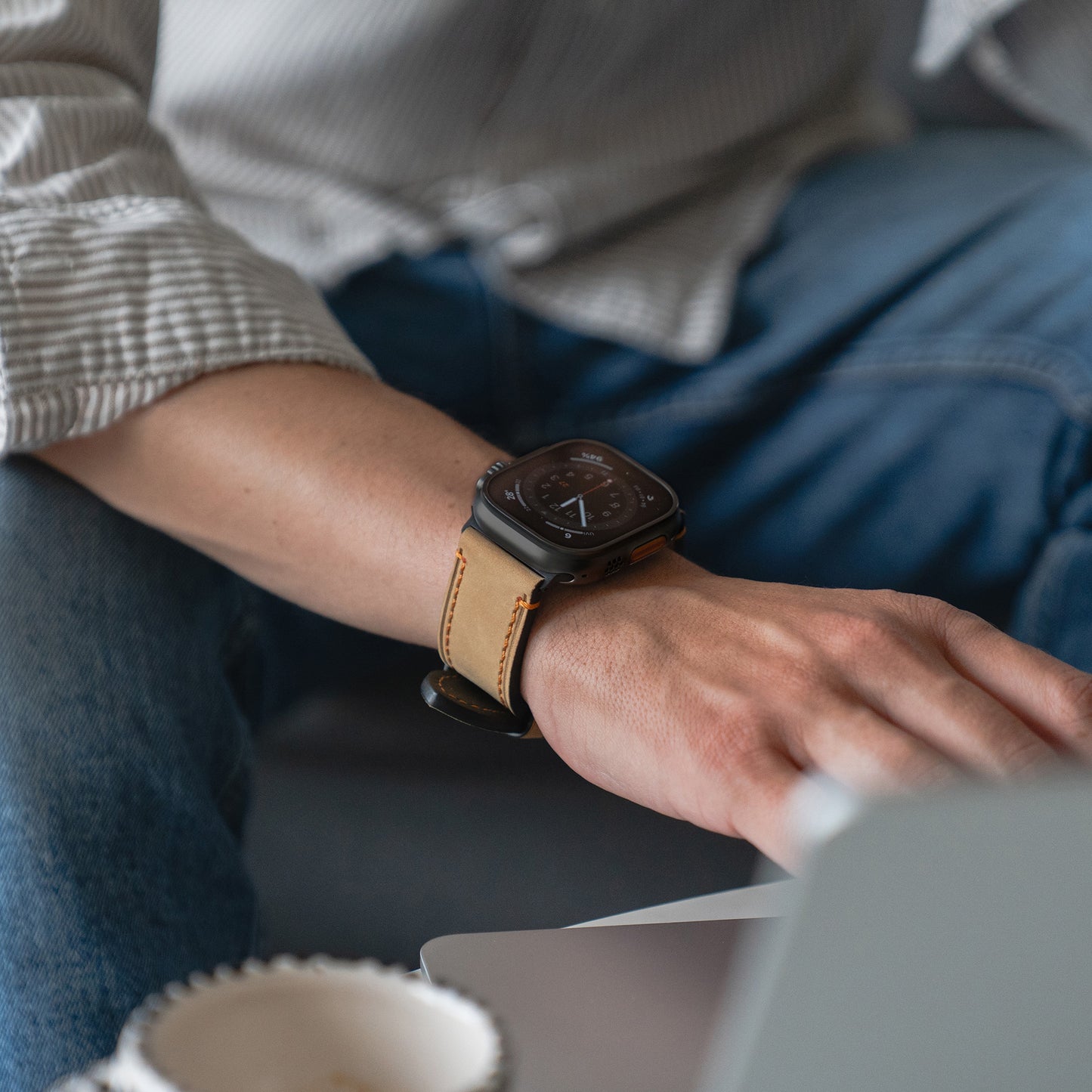 Eine Person, die ein Arktisband Apple Watch Ultra PALERMO GRANDE Lederarmband trägt, sitzt neben einem Laptop und einer Tasse.