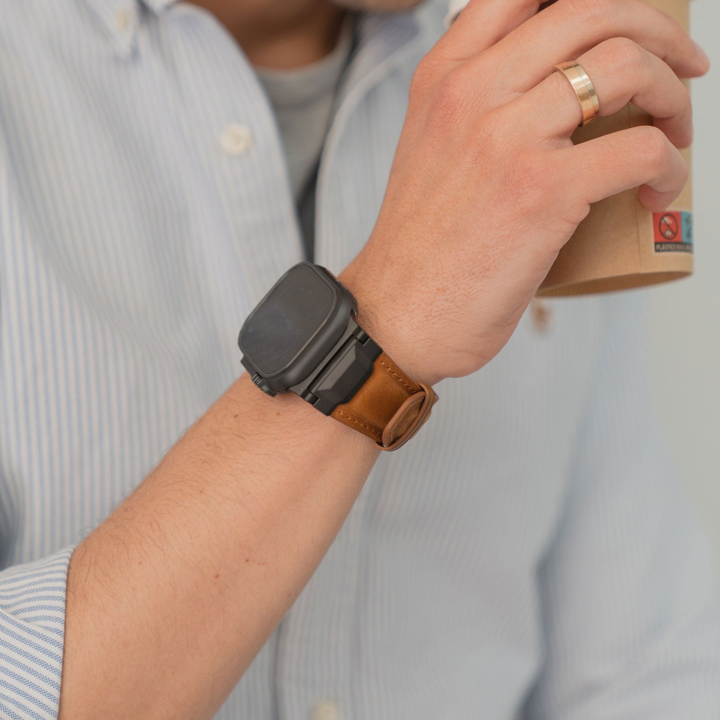 Eine Person im gestreiften Hemd hält einen Pappbecher und trägt ein Arktisband Apple Watch Ultra Lederarmband "Delta" mit braunem Lederarmband und Edelstahl Dornschließe.