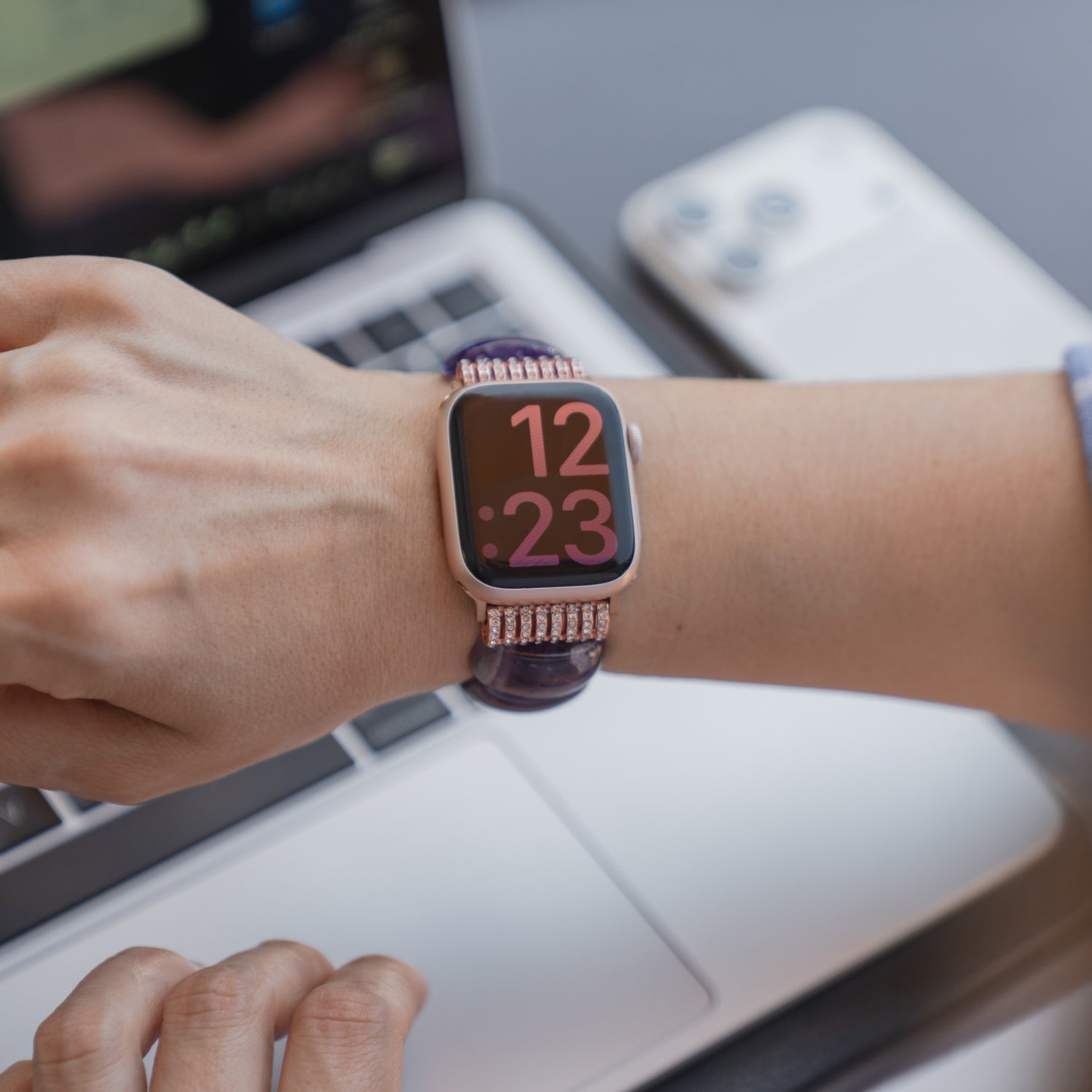Eine Person schaut auf das Arktisband-Armband ihrer Apple Watch „Stone Pearl“, auf dem 12:23 Uhr angezeigt wird, während sie an einem Laptop arbeitet; daneben liegt ein Handy.
