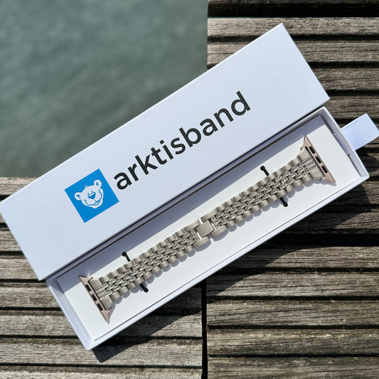 Ein arktisband Apple Watch Gliederarmband „Diamonds“ von arktisband aus silbernem Edelstahl befindet sich in einer weißen Box mit einem blauen Eisbär-Logo, platziert auf Holz in der Nähe von Wasser.