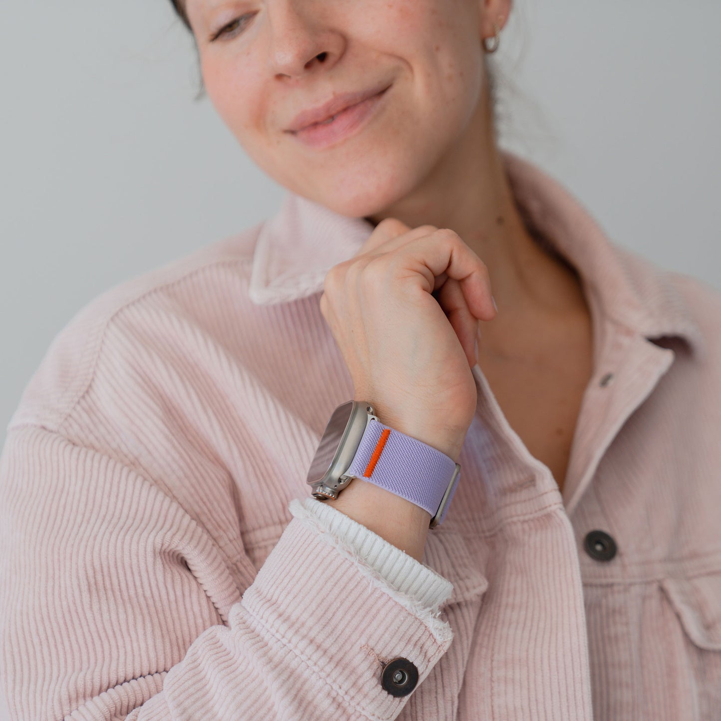 Eine Frau in einer hellrosa Cordjacke präsentiert ihr Arktisband Apple Watch Nylonarmband "FlexOne", ein lila-orangefarbenes Band, das Stil und Komfort vereint.