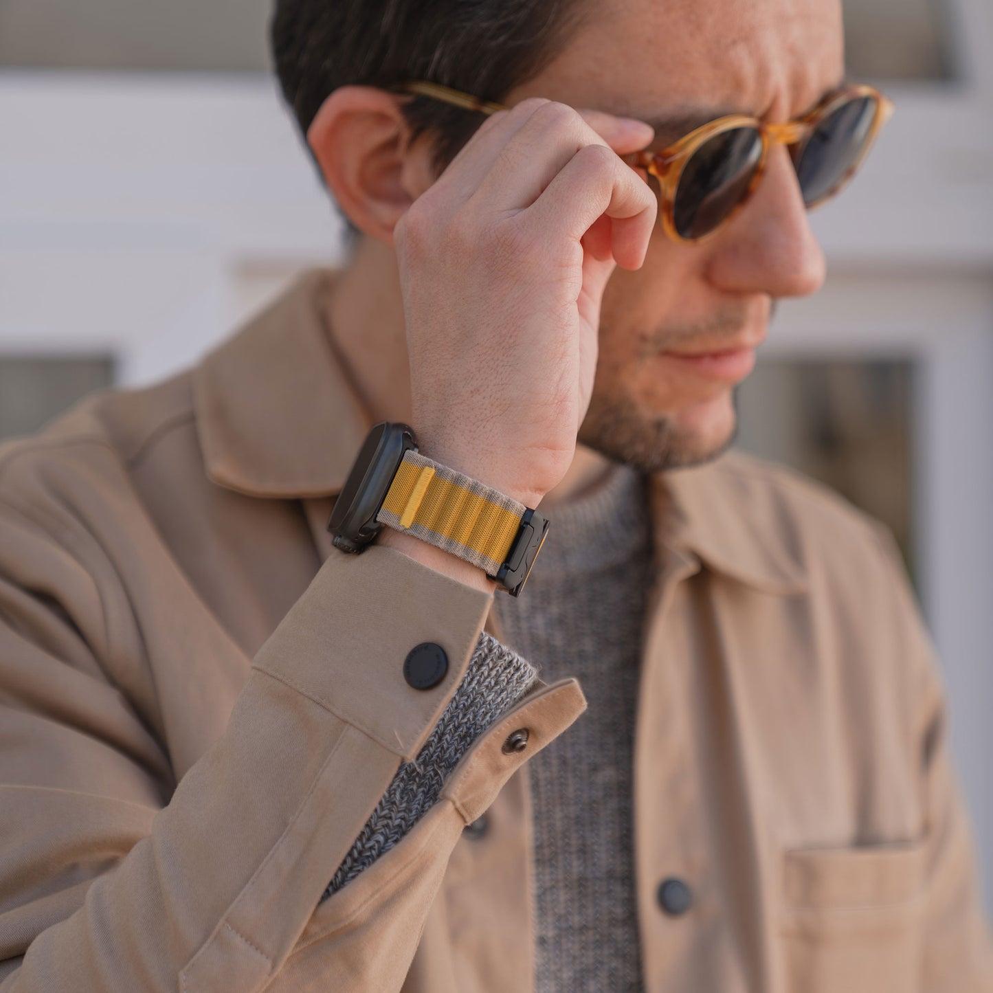 Ein Mann mit Sonnenbrille und beiger Jacke rückt seine Brille zurecht und enthüllt ein arktisband Apple Watch Armband "CoreX" mit gelb gestreiftem Wave-Look und stylischem Parachute-Verschluss.