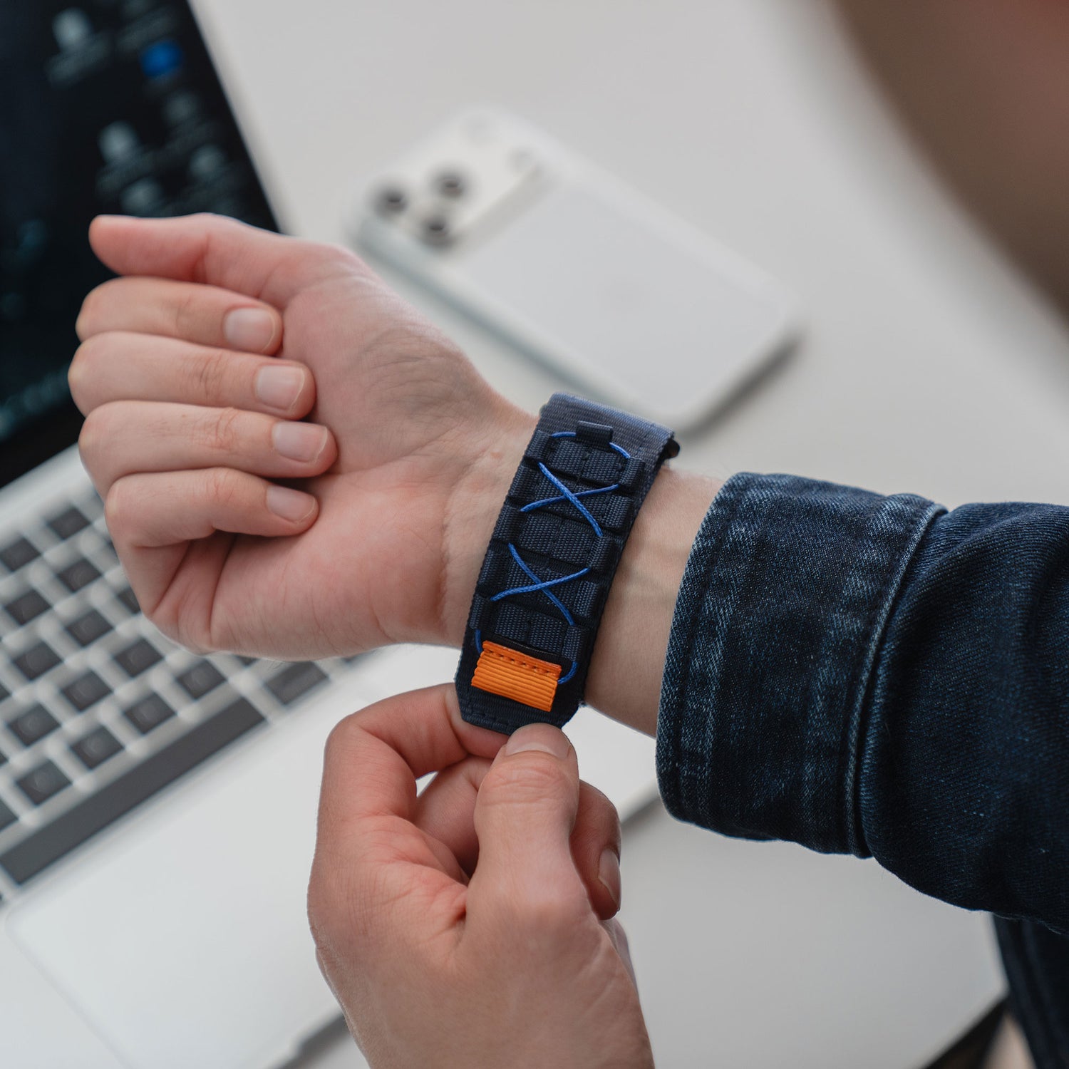 Eine Person passt das arktisband Apple Watch Outdoor-Armband "Forge" - ein blau-orangefarbenes Nylonband - an ihrem Handgelenk an, in der Nähe eines Laptops und eines Smartphones, und hebt damit dieses Outdoor-Accessoire von arktisband hervor.