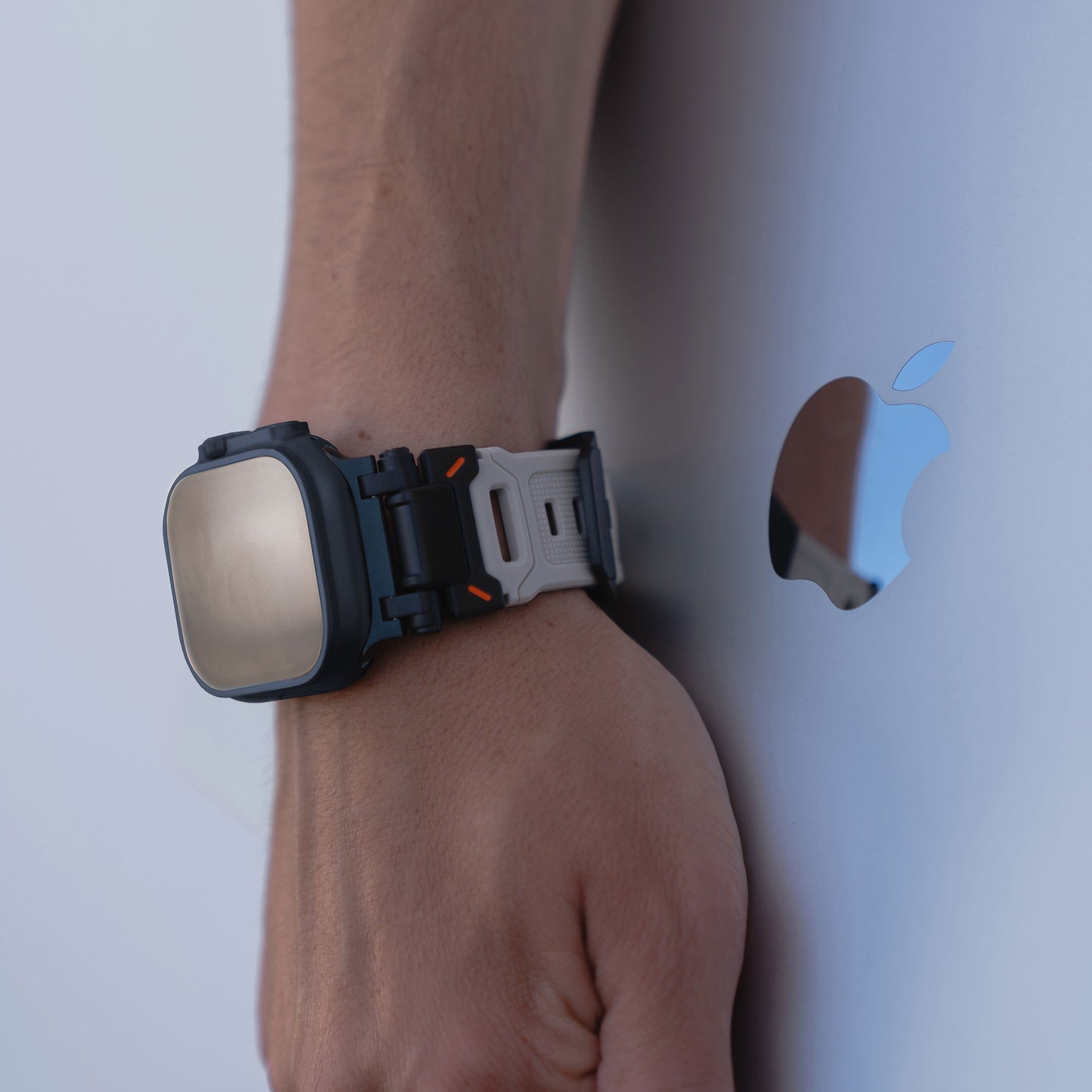 Am Handgelenk trägt man das arktisband Apple Watch Ultra Armband „Atlas“ von arktisband mit orangefarbenen Akzenten, das auf einer metallischen Oberfläche mit reflektierendem Apple-Logo ruht.
