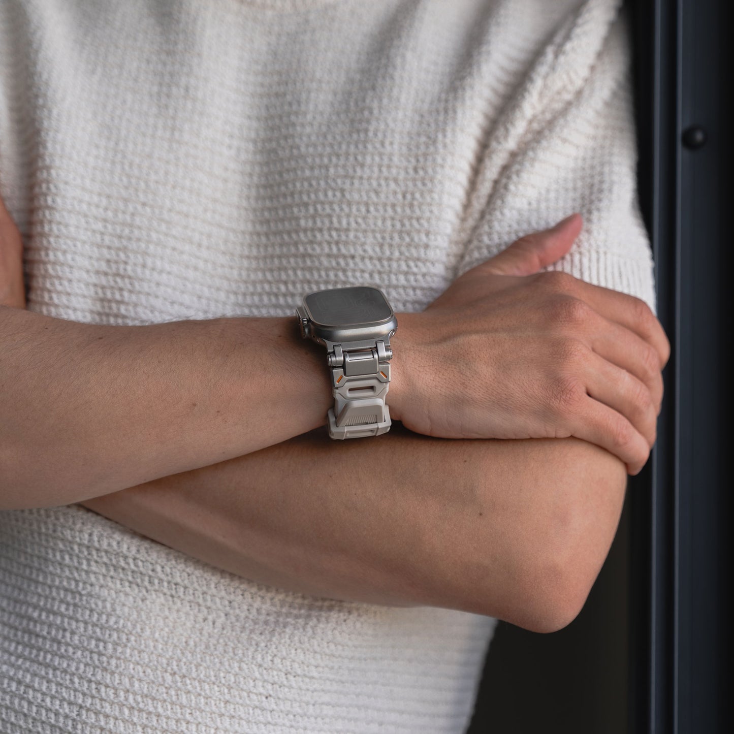 Eine Person in einem strukturierten cremefarbenen Kurzarmhemd steht mit verschränkten Armen da und präsentiert das Arktisband Apple Watch Ultra Armband „Atlas“ an ihrem Handgelenk. Der Hintergrund erscheint unscharf.