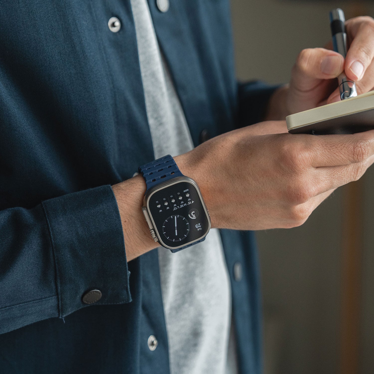 Eine Person trägt ein dunkelblaues magnetisches Silikonarmband „Power“ von Arktis für die Apple Watch am linken Handgelenk und schreibt in ein kleines Notizbuch. Sie trägt ein dunkelblaues Hemd mit Knöpfen über einem grauen T-Shirt.