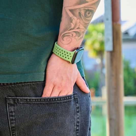 Eine Person mit Unterarm-Tattoo trägt das grüne arktisband Apple Watch Silikonarmband „Nova“ von arktisband, kombiniert mit dunklen Jeans. Sie steht im Freien, eine Hand in der Gesäßtasche; der unscharfe Hintergrund unterstreicht den stylischen Tragekomfort.