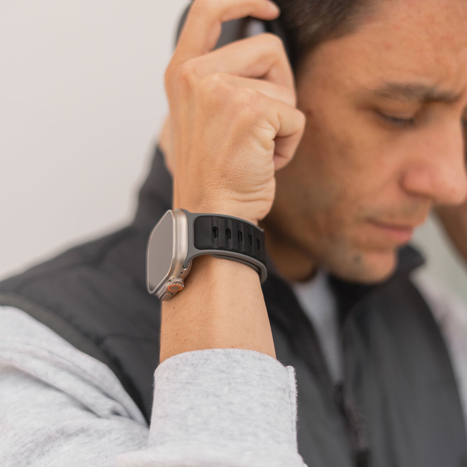 Eine Person, die ein grau-schwarzes Arktisband-Silikonarmband „Swift“ für die Apple Watch trägt, rückt Kopfhörer zurecht, während sie nach unten blickt. Sie ist mit einem grauen Sweatshirt und einer schwarzen Weste bekleidet.