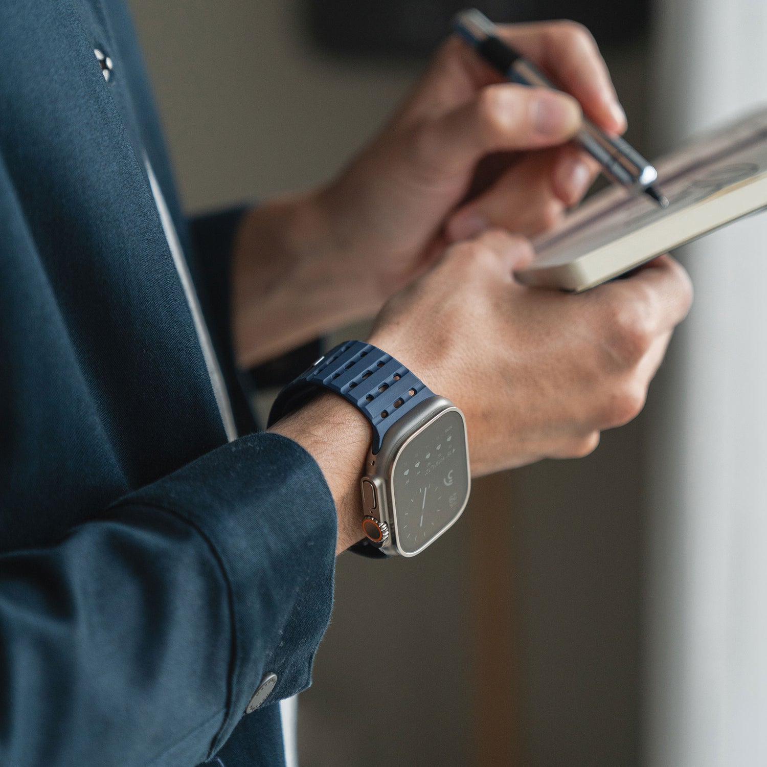 Eine Person trägt ein arktisband Apple Watch magnetisches Silikonarmband „Power“ mit blauem Silikon und Magnetverschluss und schreibt in ein Notizbuch. Sie trägt eine dunkle Jacke und ein helles Hemd; das Armband ist Apple Watch kompatibel von arktis.