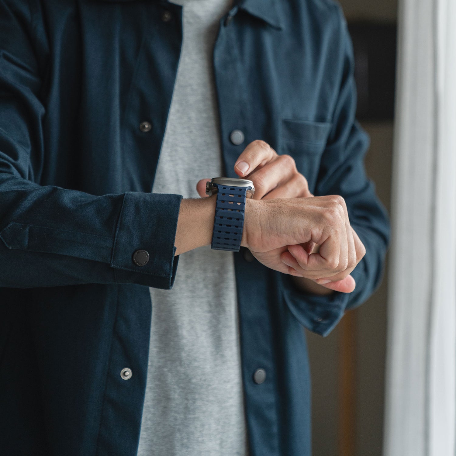 Eine Person in einem dunkelblauen Hemd über einem grauen T-Shirt rückt ein magnetisches Silikonarmband „Power“ von arktis für die Apple Watch an ihrem linken Handgelenk zurecht. Die Innenszene wird von rechts natürlich beleuchtet.