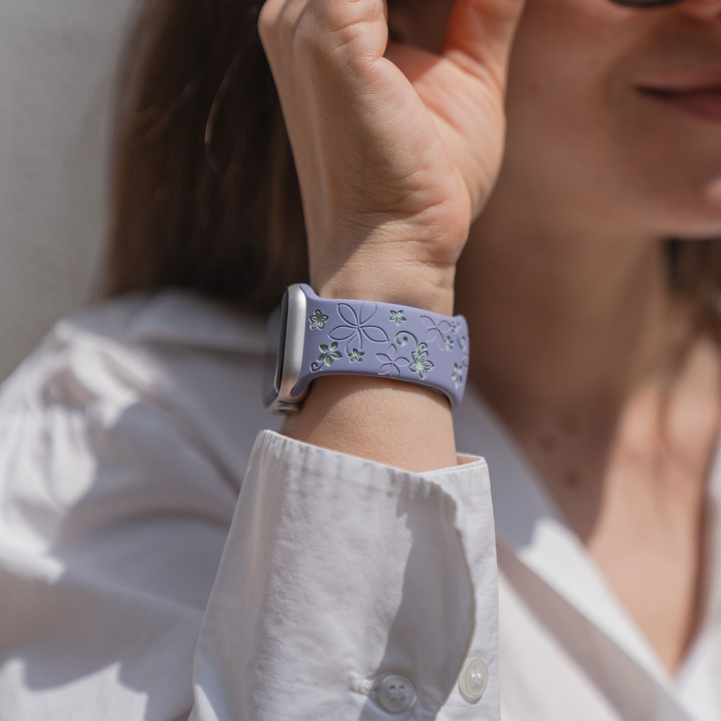 Eine Frau in einem weißen Hemd präsentiert das Arktisband Apple Watch Silikonarmband "Bloom" in lavendelfarbenem Blumenmuster und hält ihre Hand an ihr Gesicht.