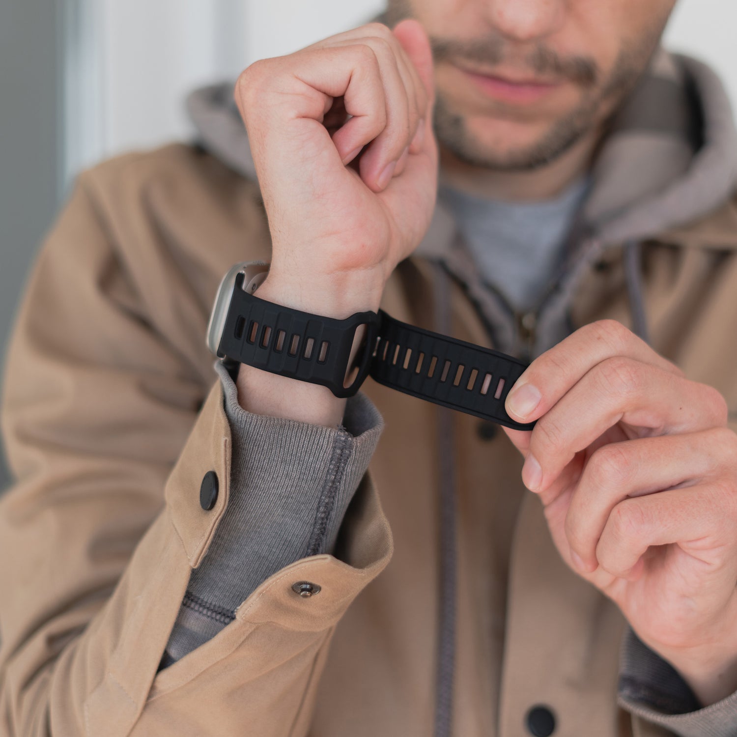 Eine Person in einer hellbraunen Jacke und einem grauen Hoodie befestigt ein Arktisband Apple Watch Silikonarmband „Runner“ um ihr Handgelenk. Das Bild hebt die atmungsaktive Bandstruktur hervor und konzentriert sich auf Hände und Handgelenk, wobei das Gesicht teilweise außerhalb des Bildes liegt.