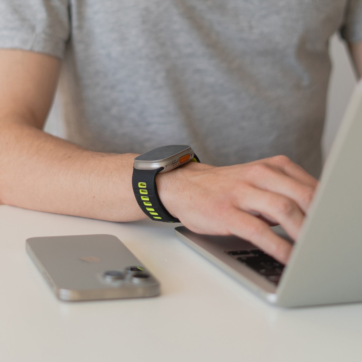 Eine Person in einem grauen T-Shirt und einem Arktisband Apple Watch Silikonarmband „Runner“ mit atmungsaktivem Design tippt auf einem Laptop. Ein Smartphone liegt mit der Vorderseite nach oben auf dem weißen Tisch daneben.