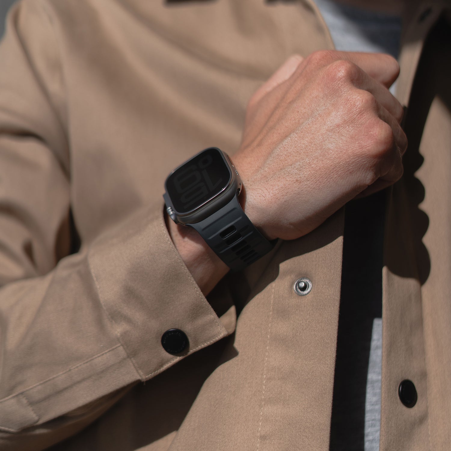 Eine Person in einer hellbraunen Jacke hält es nahe ihrer Brust und zeigt das Zifferblatt eines Apple Watch-kompatiblen Arktisband-Silikonarmbands „Runner“ in dunkler Farbe.
