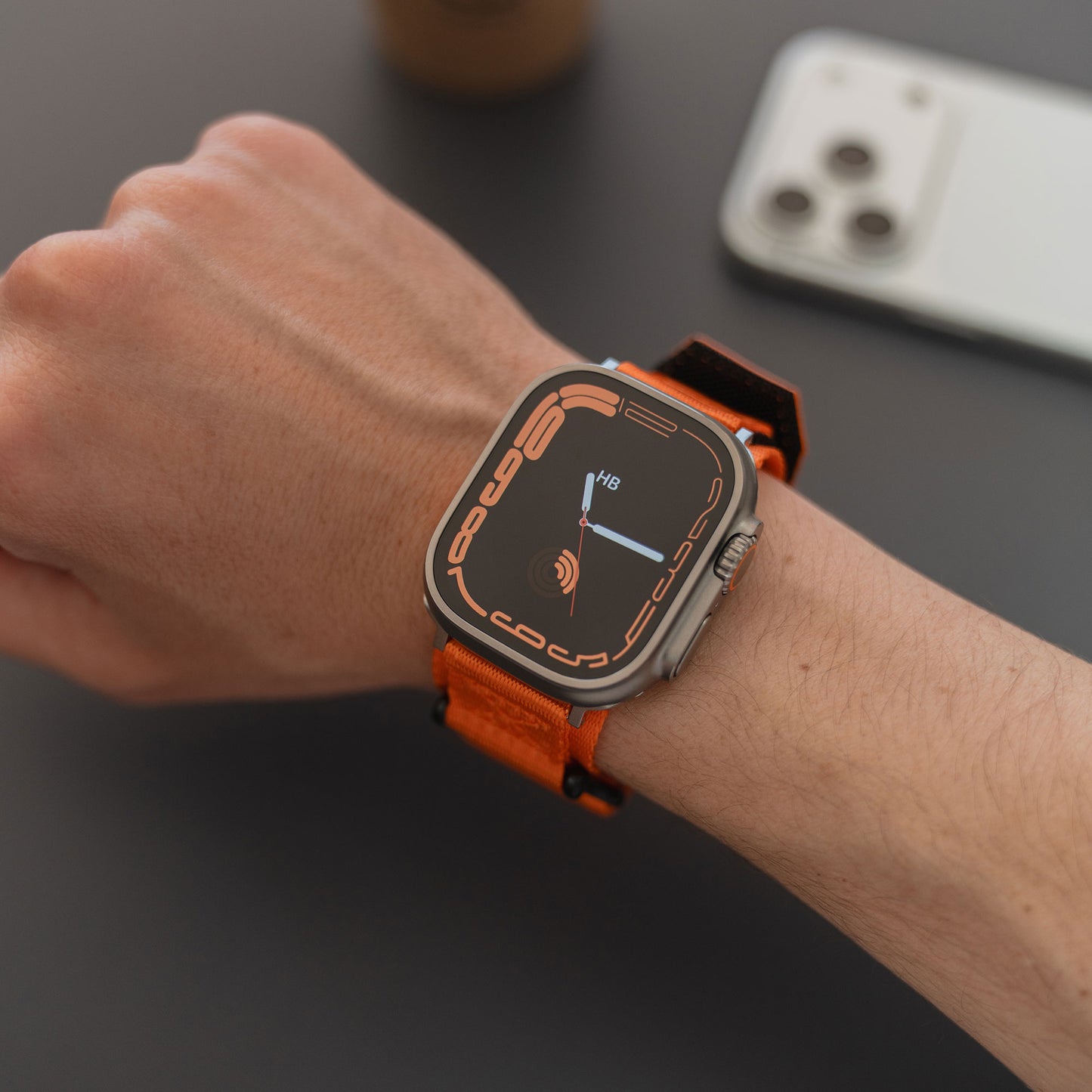 Eine Hand, die das arktisband Apple Watch Outdoor Armband "Forge" mit orangefarbenem Nylonband trägt, ruht neben einem weißen Smartphone auf einer dunklen Fläche.
