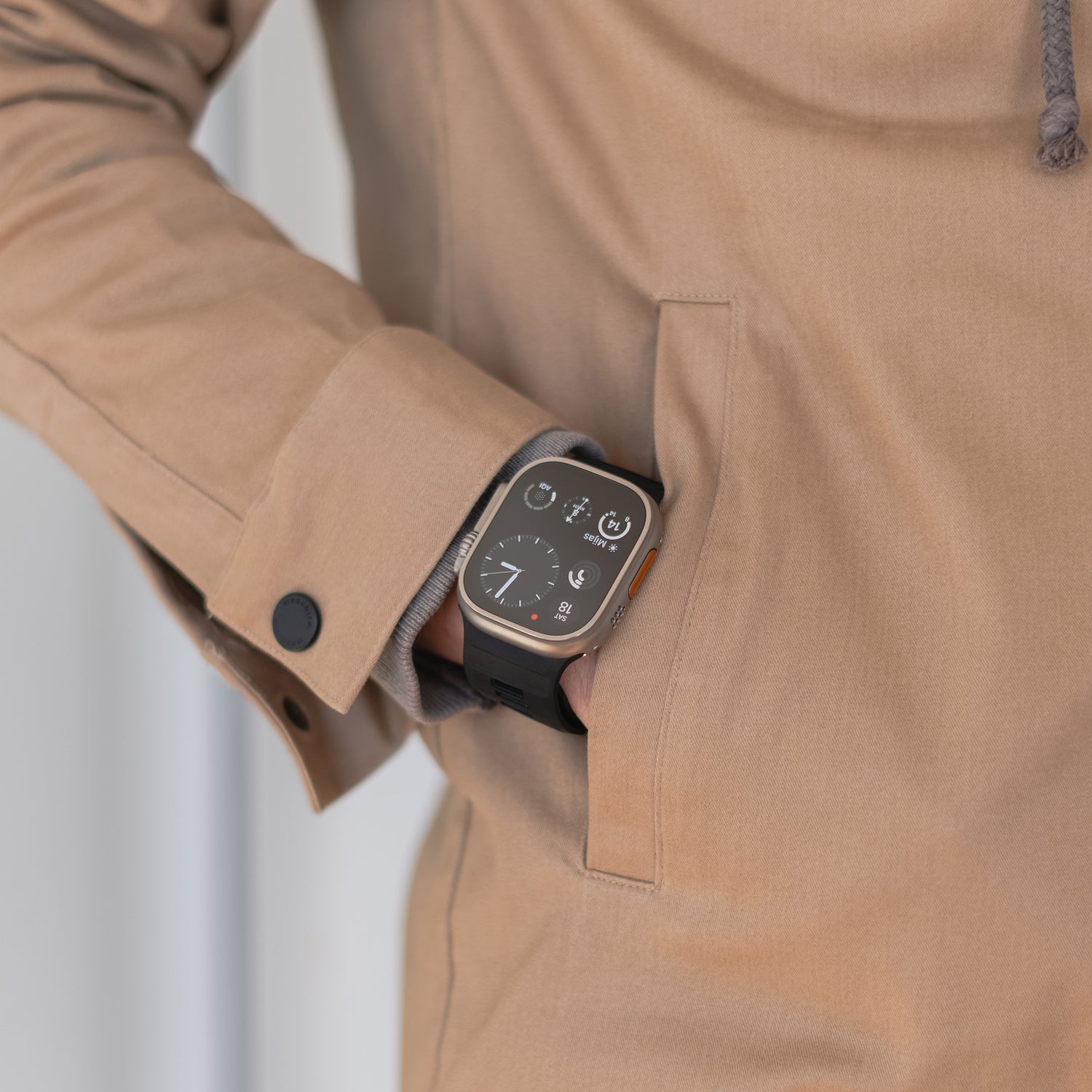 Eine Person in einer beigen Jacke, deren linke Hand in der Tasche steckt, trägt ein Arktisband Apple Watch Silikonarmband „Runner“ – das schwarze, atmungsaktive Armband ist auf ihrer Smartwatch zu sehen, da auf dem quadratischen Zifferblatt Uhrzeit und Symbole angezeigt werden.