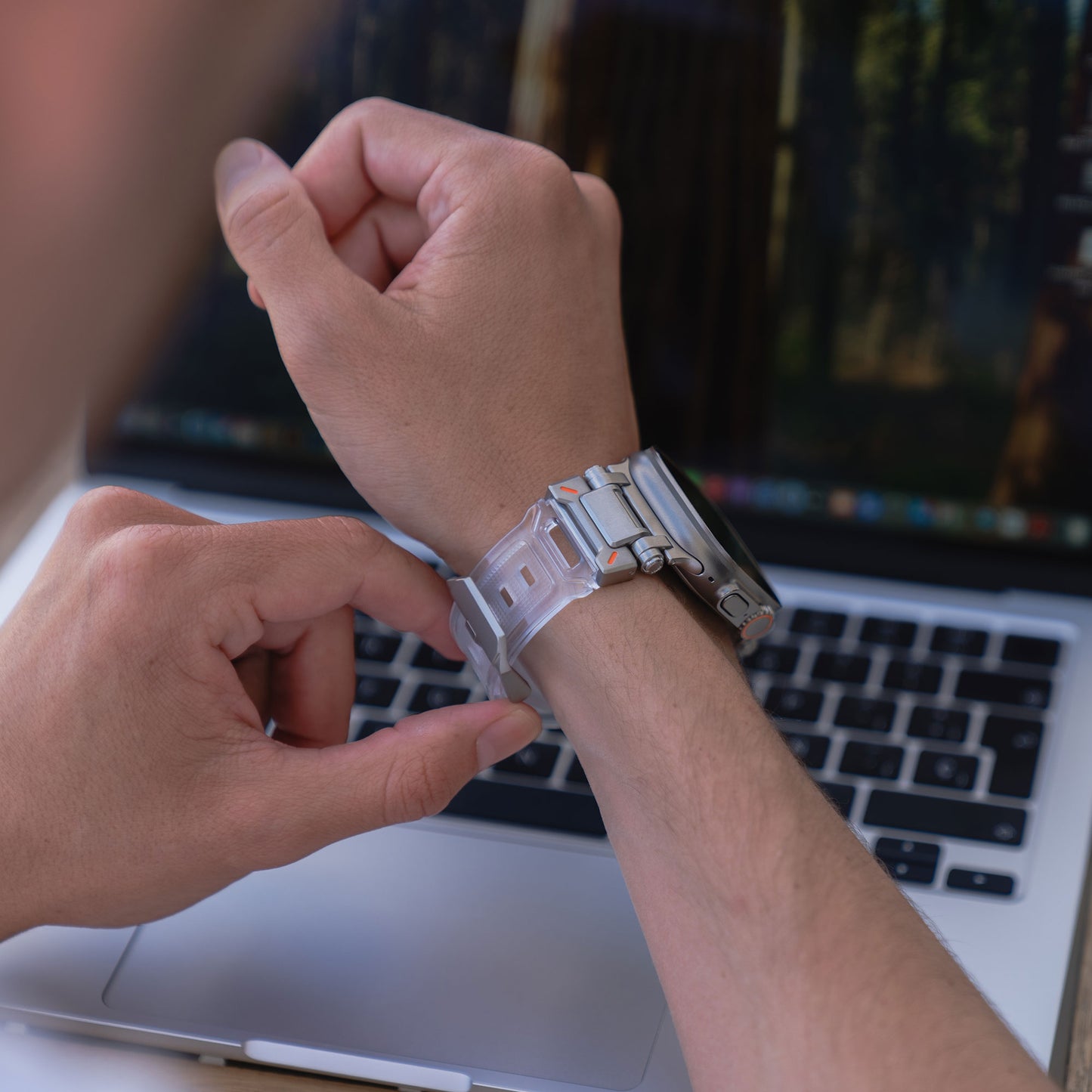 Eine Person passt das aus TPU und Edelstahl gefertigte Arktisband Apple Watch Ultra Armband „Atlas“ an, während sie vor einem geöffneten Laptop sitzt.