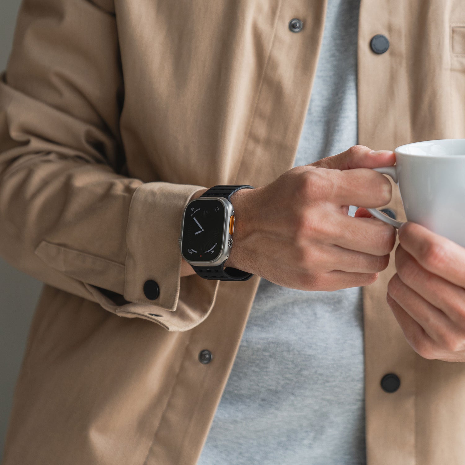 Eine Person in beiger Jacke und grauem Hemd hält eine weiße Kaffeetasse und trägt am anderen Handgelenk ein magnetisches Silikonarmband „Power“ von arktis für die Apple Watch.