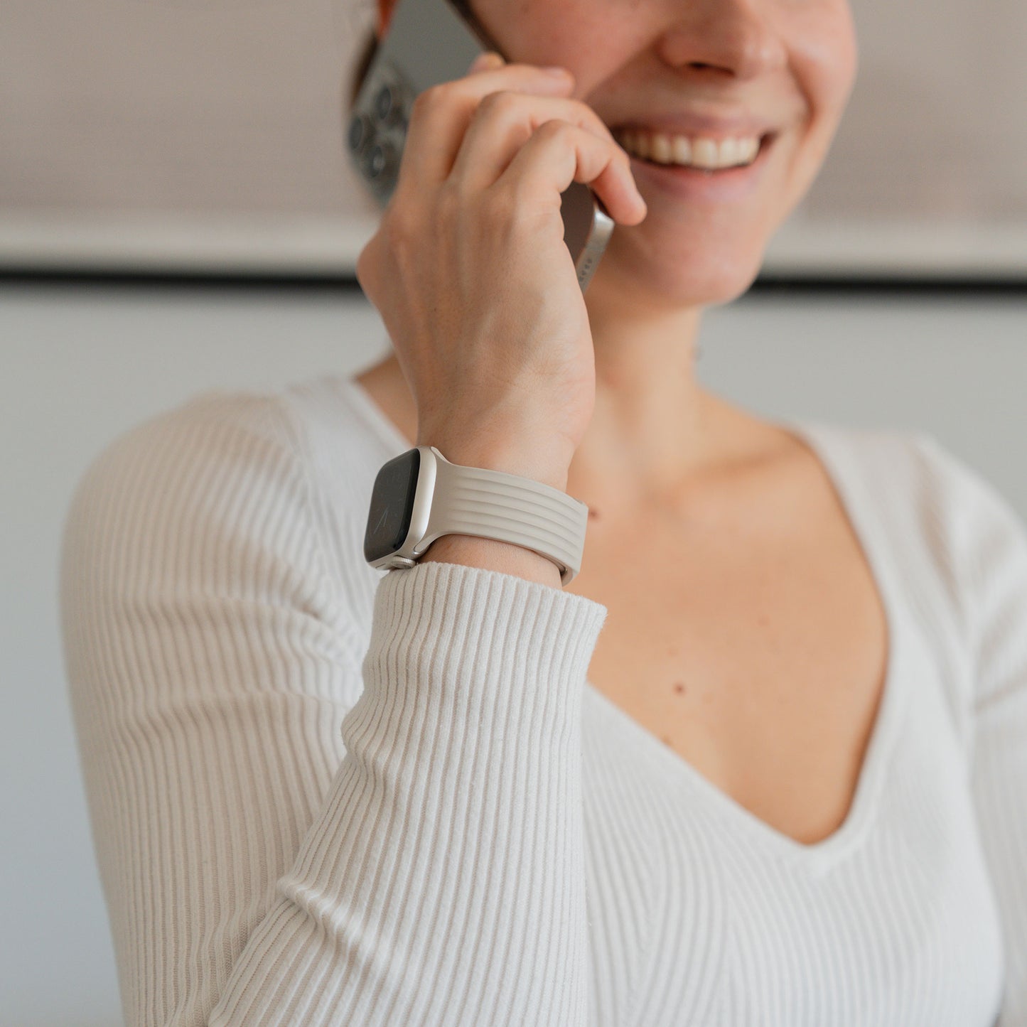 Eine lächelnde Frau in einem weißen Top telefoniert, während sie das arktisband Apple Watch Armband "Skandi" von arktisband aus weichem Fluorelastomer am Handgelenk trägt.