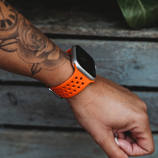 Eine Person mit einem Rosentattoo auf dem Unterarm trägt ein Arktisband Apple Watch Silikonarmband „Nova“ in Orange und hält ihr Handgelenk über einer verwitterten Holzoberfläche mit grünen Blättern im Hintergrund.