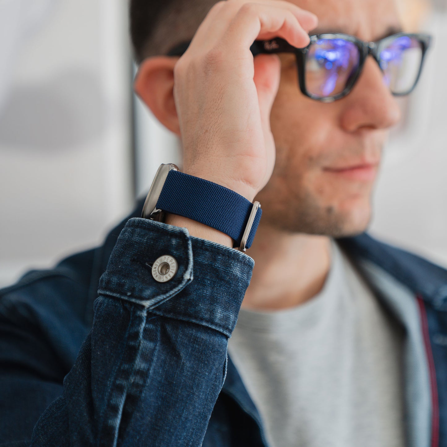 Mann mit Brille, Jeansjacke und Apple Watch Nylonarmband "FlexOne" mit blauem Armband, in einer unauffälligen Umgebung.