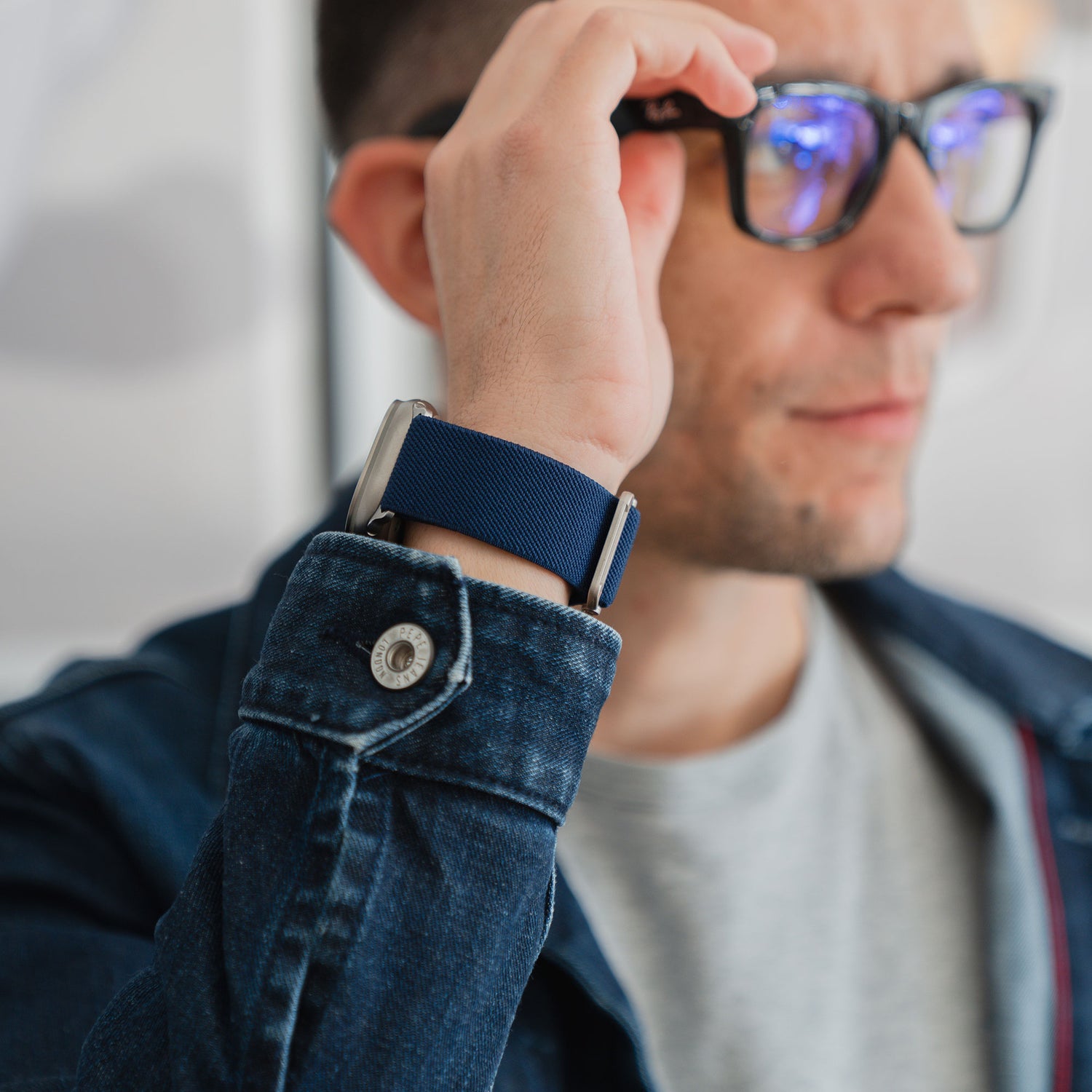 Mann mit Brille, Jeansjacke und Apple Watch Nylonarmband "FlexOne" mit blauem Armband, in einer unauffälligen Umgebung.