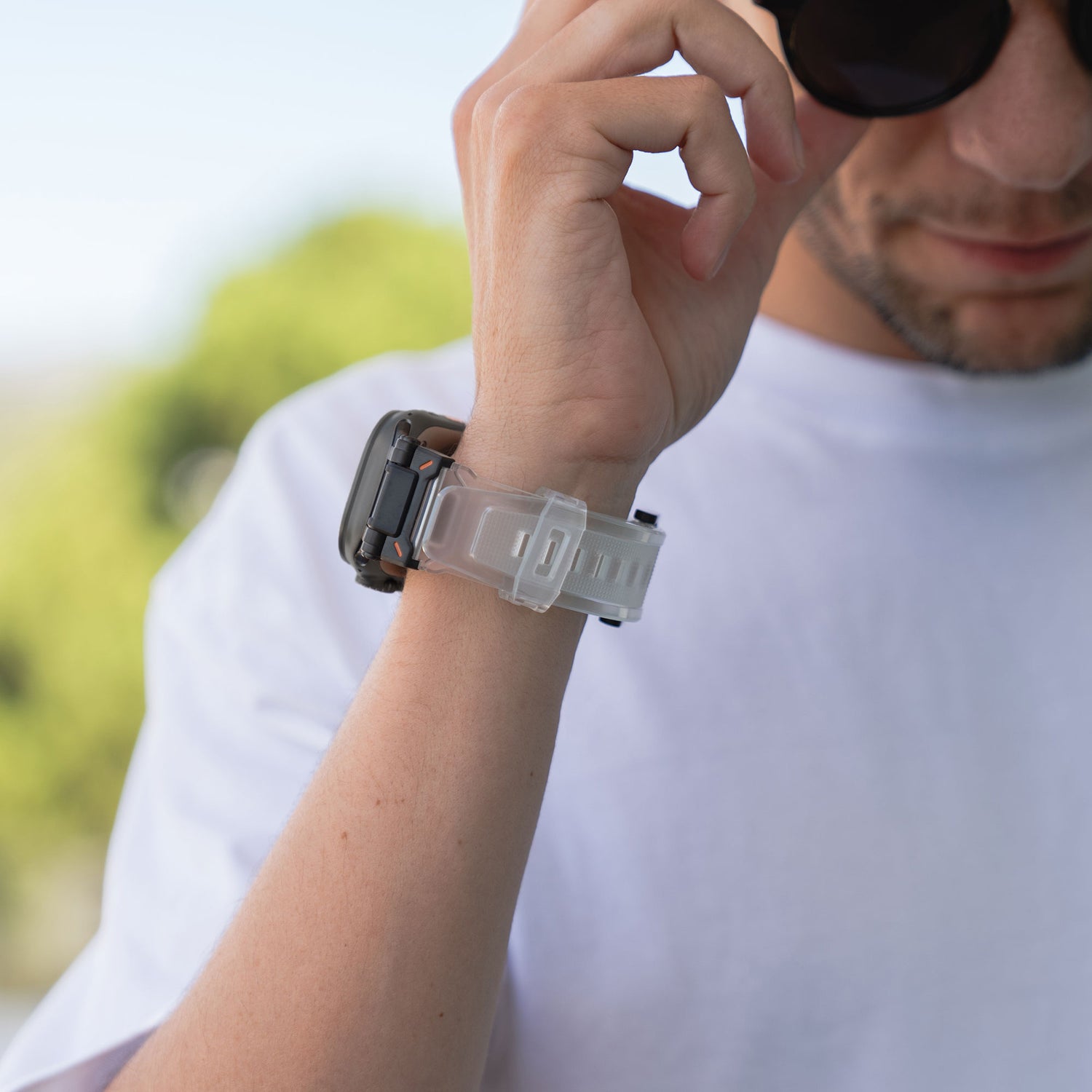 Eine Person im weißen T-Shirt rückt ihre Sonnenbrille zurecht und enthüllt ein Arktisband Apple Watch Ultra Armband „Atlas“ an ihrem Handgelenk. Im Hintergrund ist weiches Grün zu sehen.