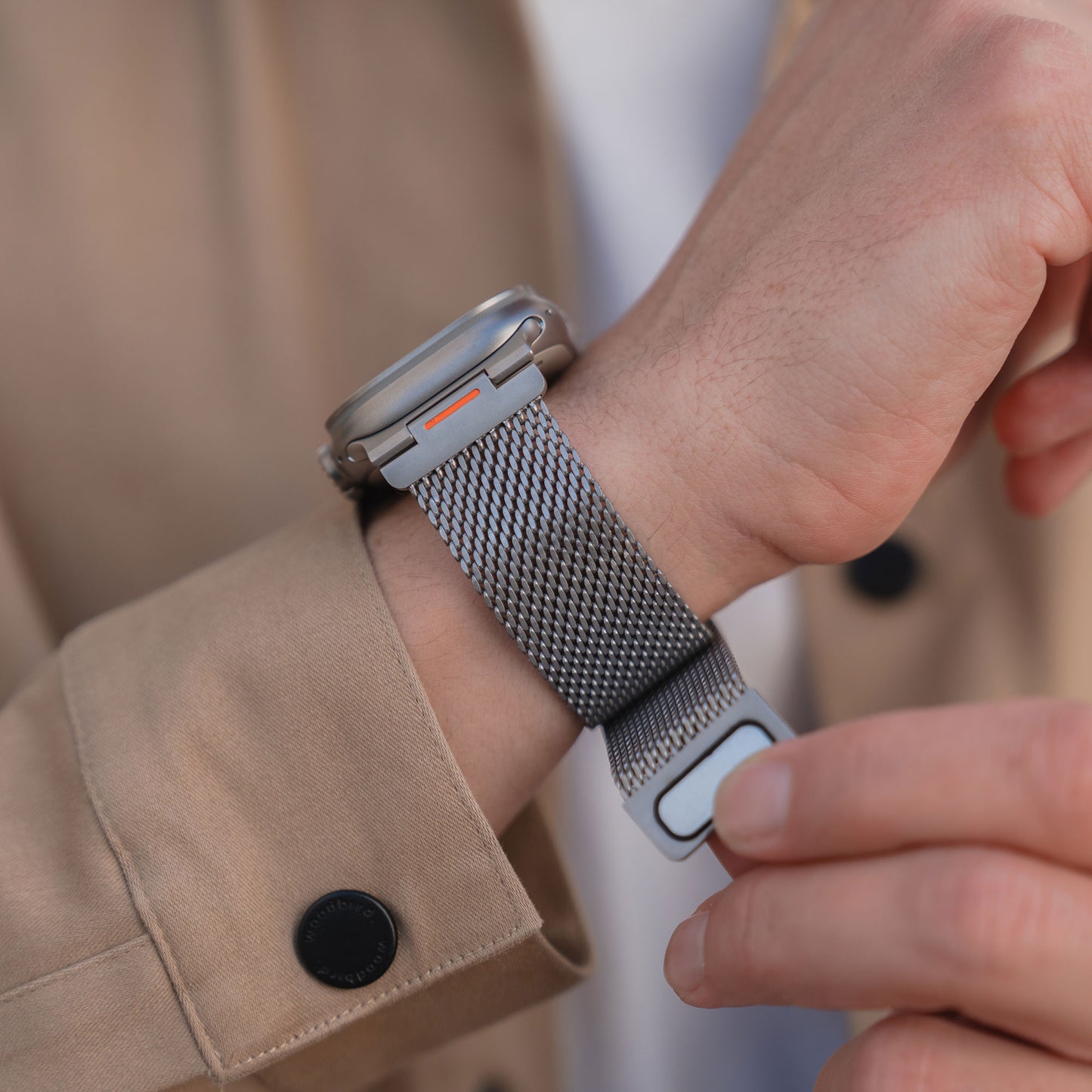 Nahaufnahme einer Person, die das Arktisband Apple Watch Milanaise Armband "Bold SE" mit Magnetverschluss an ihrem Handgelenk befestigt, gestylt mit einem beigen Mantel.