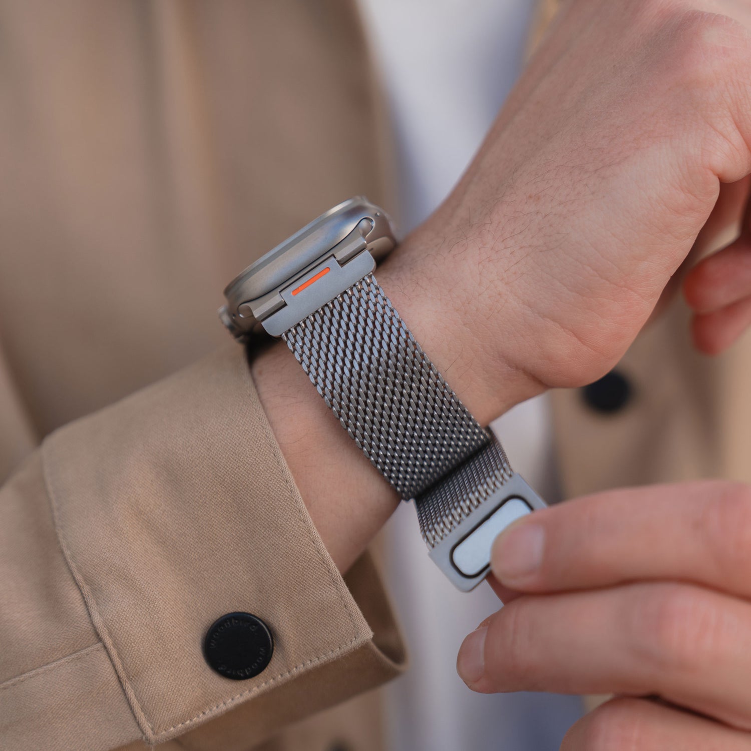 Nahaufnahme einer Person, die das Arktisband Apple Watch Milanaise Armband "Bold SE" mit Magnetverschluss an ihrem Handgelenk befestigt, gestylt mit einem beigen Mantel.