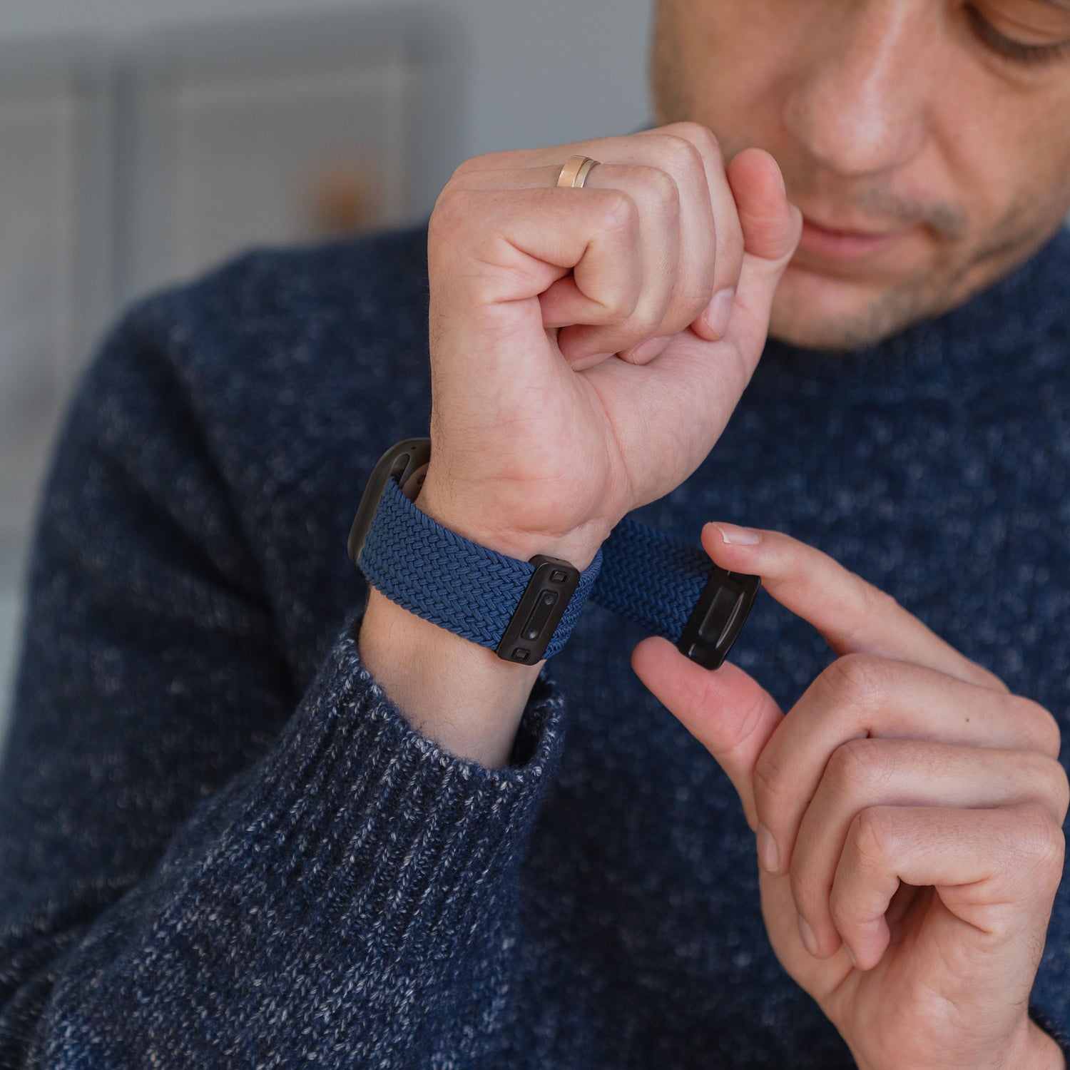 Eine Person, die sich das Arktisband „Geflochtenes Apple Watch Ultra Armband ‚Casual‘“ mit Magnetverschluss in Blau am Handgelenk befestigt, während sie einen blauen Pullover und einen goldenen Ring trägt.