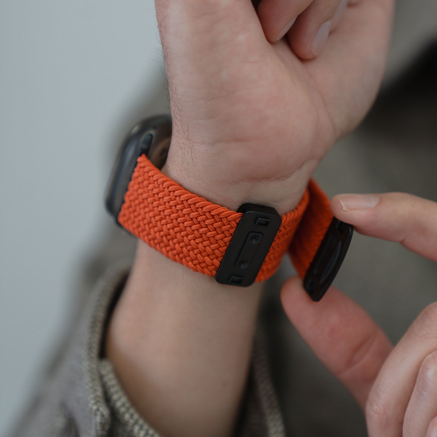 Eine Person befestigt mit beiden Händen ein orangefarbenes Arktisband – ein geflochtenes Apple Watch Ultra-Armband „Casual“ mit Magnetverschluss – am Handgelenk.
