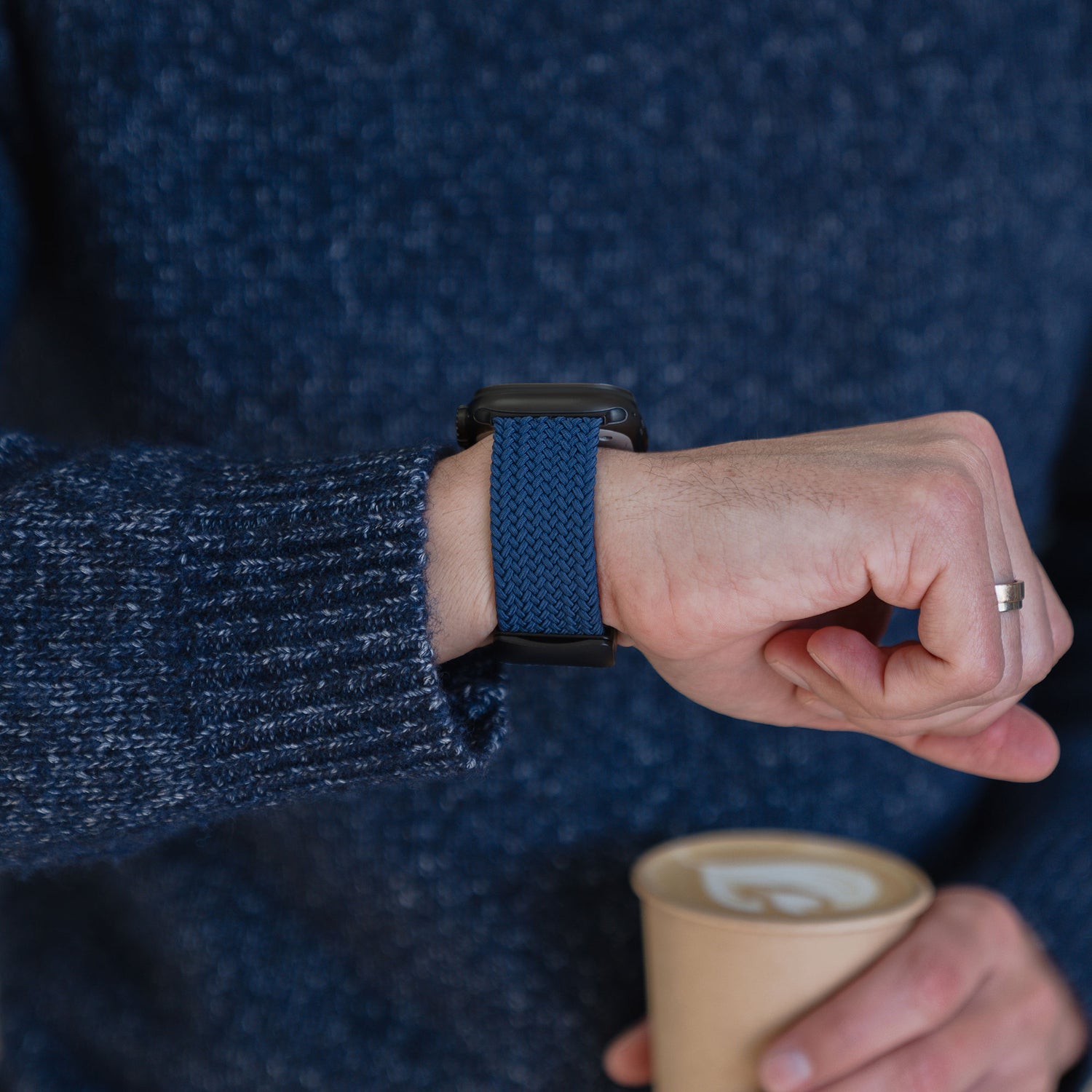 Eine Person, die das „Geflochtenes Apple Watch Ultra Armband ‚Casual‘“ mit Magnetverschluss von arktisband in Blau trägt, eine Tasse Kaffee in der Hand hält und einen blauen Pullover trägt.