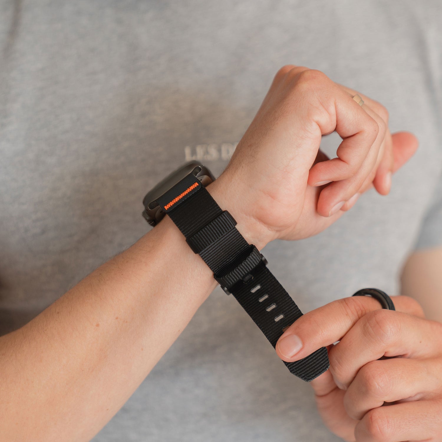 Eine Person trägt ein graues T-Shirt und befestigt das arktisband Apple Watch Armband "Terra" von arktisband, mit seinem 24 mm extrabreiten schwarzen Outdoor-Nylon-Armband, an ihrem linken Handgelenk.