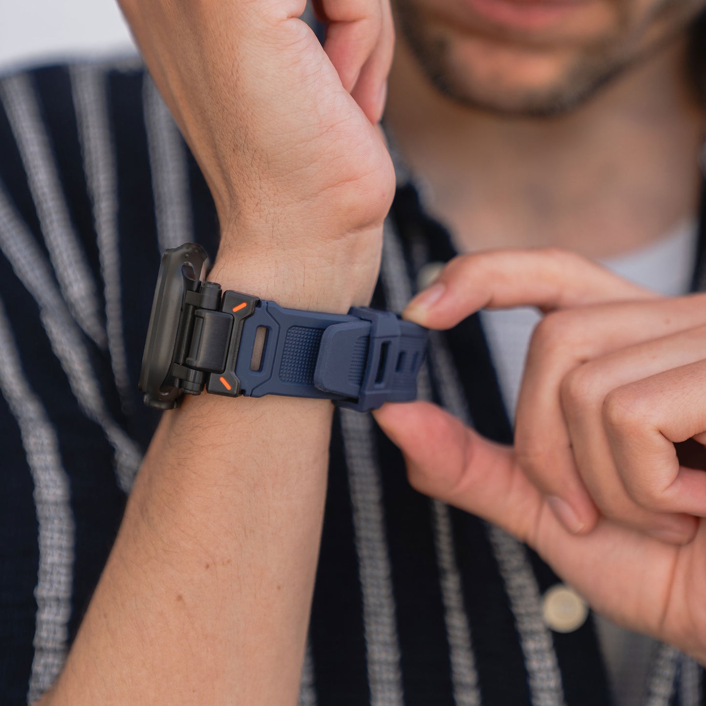 Eine Person im gestreiften Hemd befestigt mit der rechten Hand das Arktisband Apple Watch Ultra Armband „Atlas“ mit Edelstahl-Elementen an ihrer schwarzen Apple Watch Ultra am linken Handgelenk.