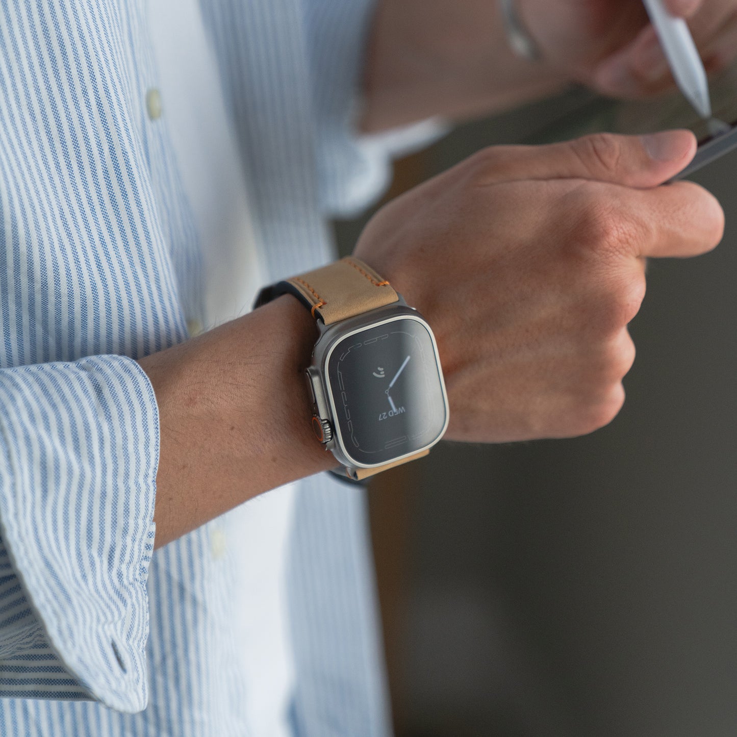 Eine Person trägt ein Arktisband Apple Watch Ultra PALERMO GRANDE Lederarmband, benutzt ein Smartphone und ist mit einem blau gestreiften Hemd bekleidet.