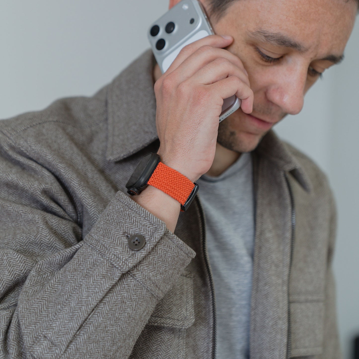 Ein Mann in einer braunen Jacke hält ein Handy ans Ohr und trägt eine Smartwatch mit dem Arktisband „Geflochtenes Apple Watch Ultra Armband ‚Casual‘“, das sich durch einen stilvollen Magnetverschluss auszeichnet.