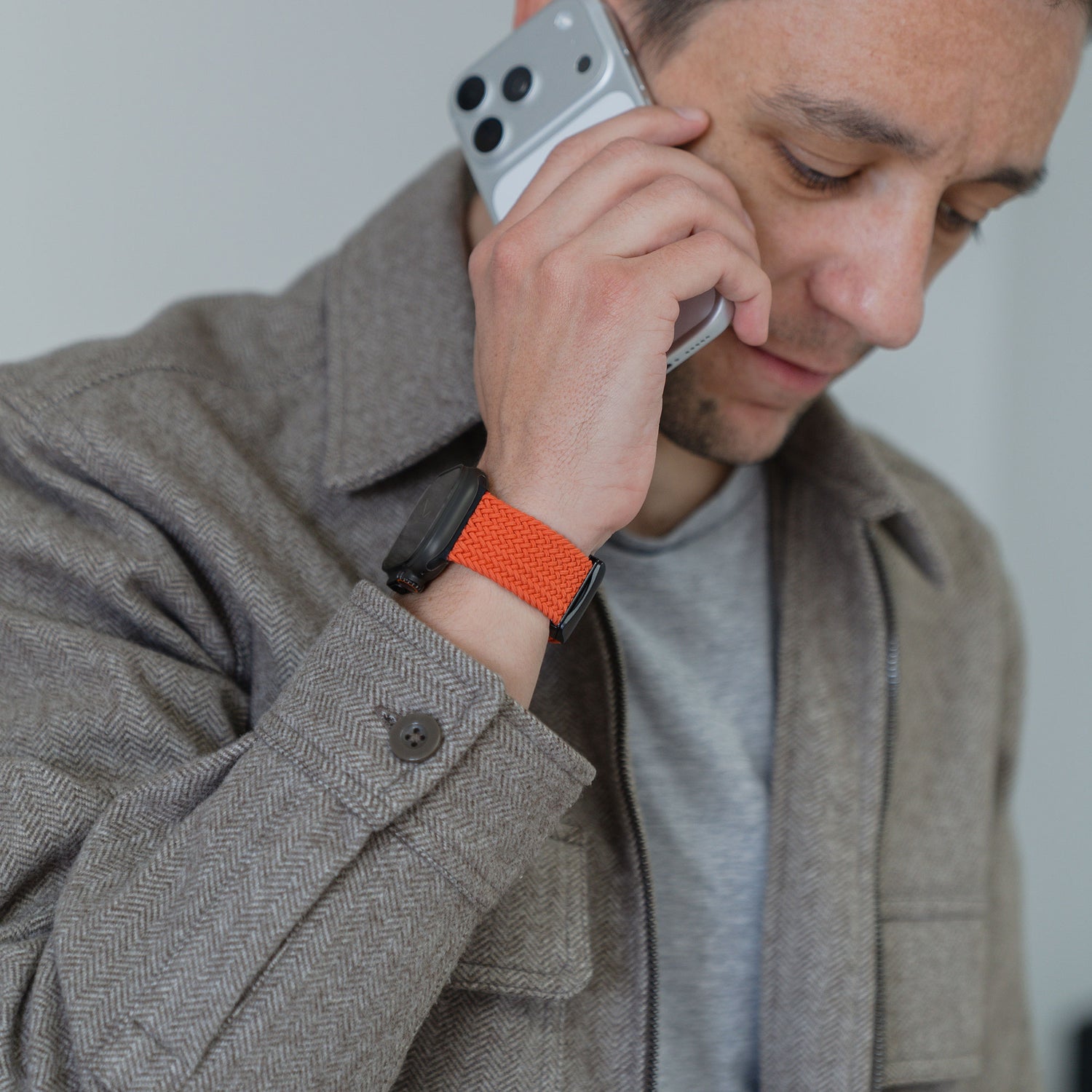 Ein Mann in einer braunen Jacke hält ein Handy ans Ohr und trägt eine Smartwatch mit dem Arktisband „Geflochtenes Apple Watch Ultra Armband ‚Casual‘“, das sich durch einen stilvollen Magnetverschluss auszeichnet.