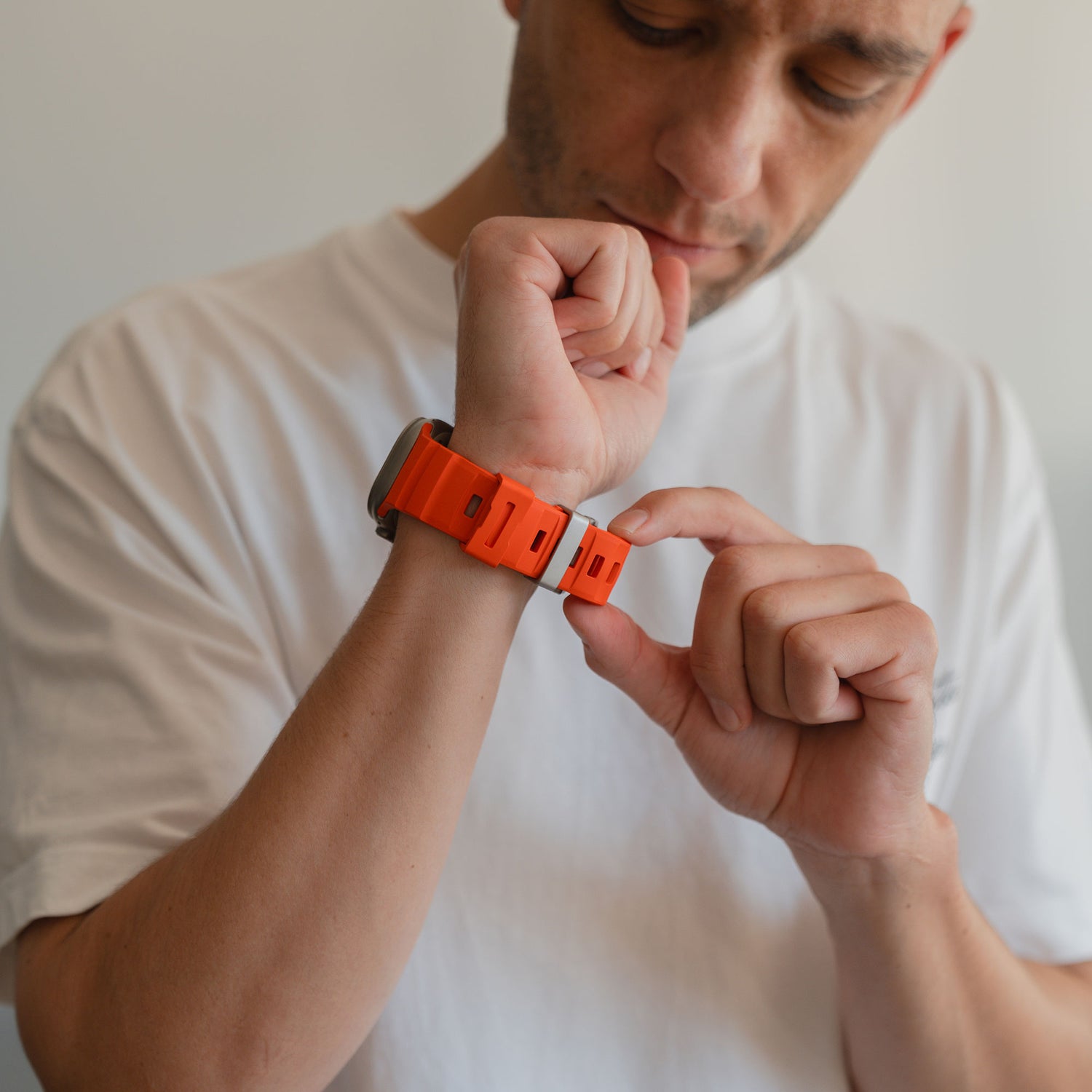 Ein Mann in einem weißen T-Shirt befestigt das orangefarbene Arktisband Apple Watch Armband "Flux" mit Edelstahl Schließe an seinem linken Handgelenk.