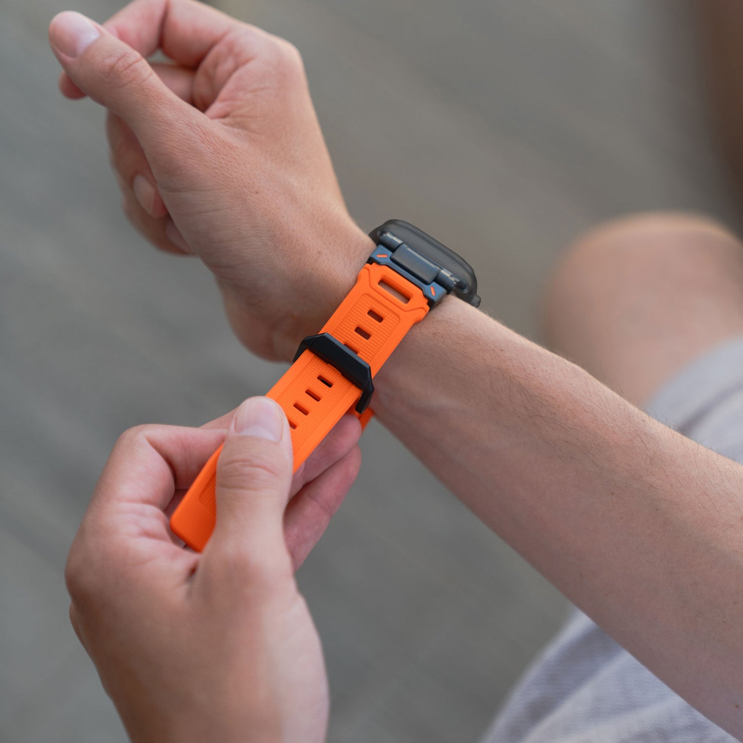 Eine Person befestigt das Arktisband Apple Watch Ultra Armband „Atlas“ in Orange mit Edelstahl-Elementen am linken Handgelenk; Uhr und robustes Armband werden hervorgehoben, während der Hintergrund unscharf wird.