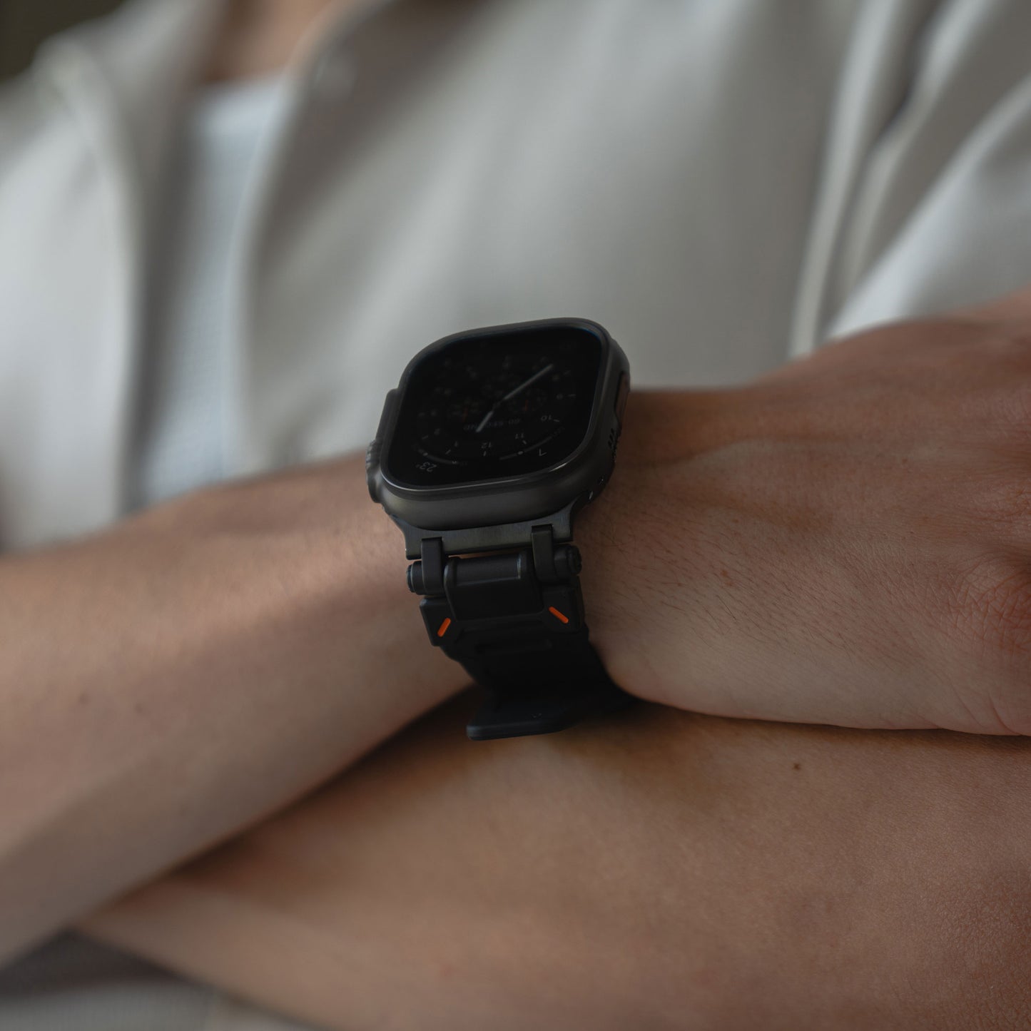 Eine Person mit verschränkten Armen trägt ein Arktisband Apple Watch Ultra Armband „Atlas“ an ihrer Apple Watch Ultra, mit quadratischem Ziffernblatt. Sie trägt ein helles Hemd über einem weißen Oberteil.