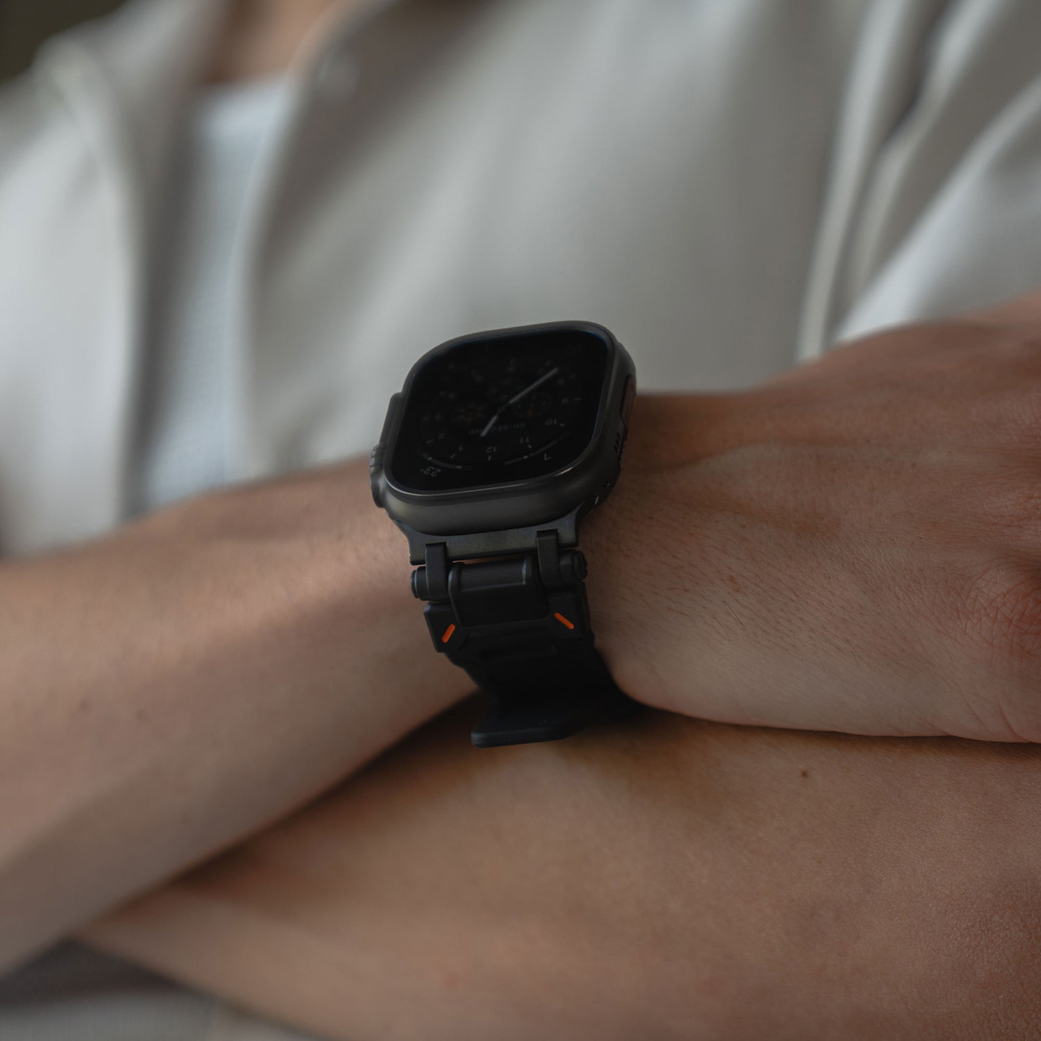 Eine Person mit verschränkten Armen trägt ein Arktisband Apple Watch Ultra Armband „Atlas“ an ihrer Apple Watch Ultra, mit quadratischem Ziffernblatt. Sie trägt ein helles Hemd über einem weißen Oberteil.