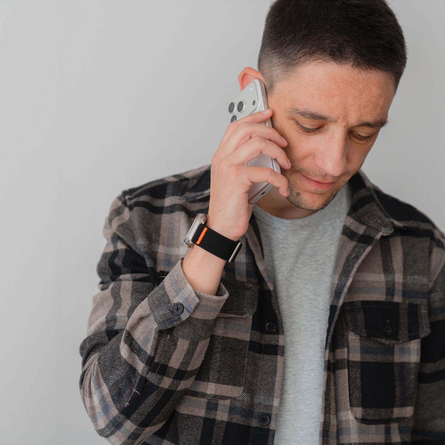 Ein Mann im karierten Hemd, der das Arktisband Apple Watch Nylonarmband "FlexOne" trägt, spricht auf seinem Smartphone und blickt mit neutraler Miene auf einen schlichten Hintergrund.