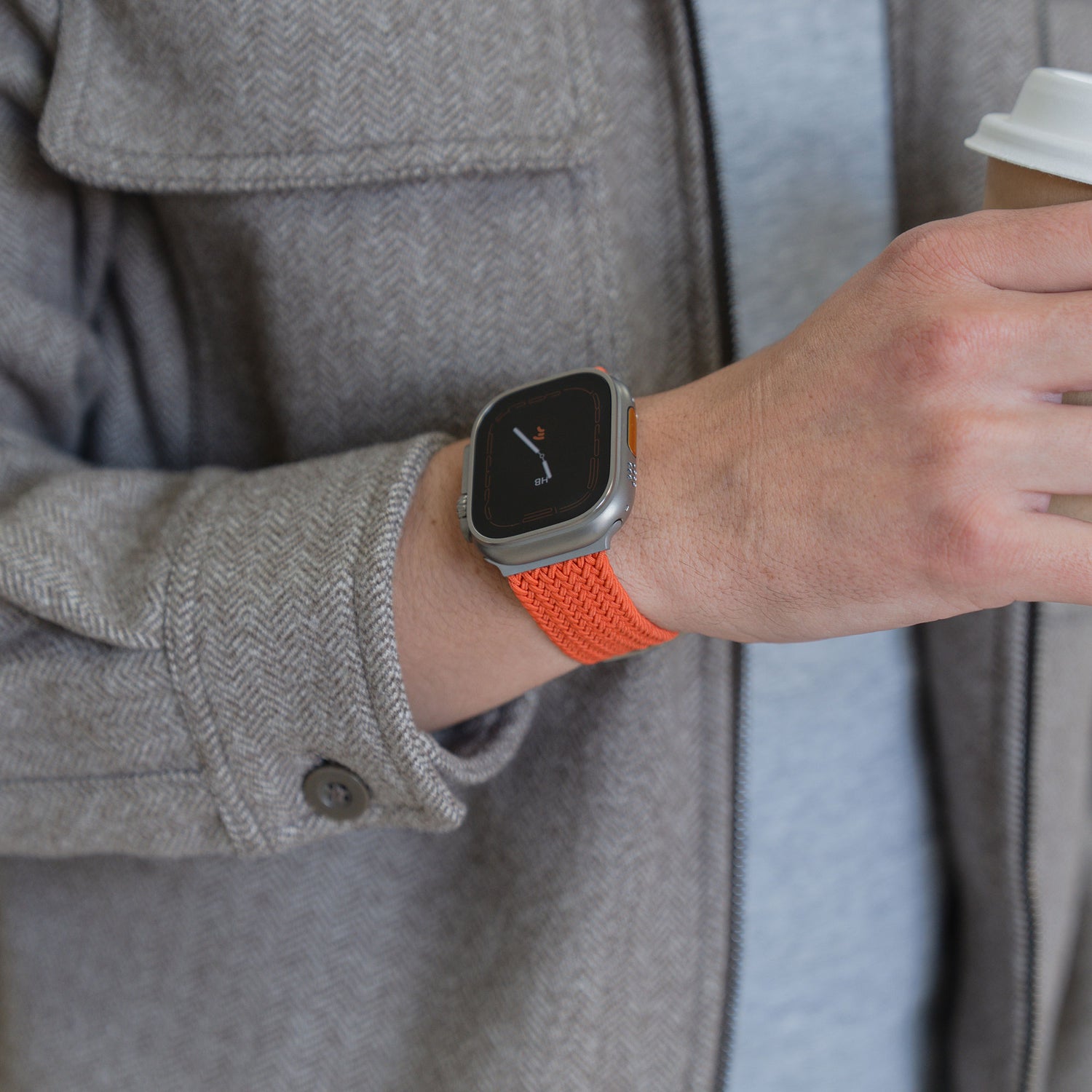 Eine Person, die ein „Geflochtenes“-Apple-Watch-Ultra-Armband „Casual“ von arktisband mit Magnetverschluss in Orange trägt, hält eine Kaffeetasse in der Hand und ist mit einer grauen Jacke und einem grauen Hemd bekleidet.
