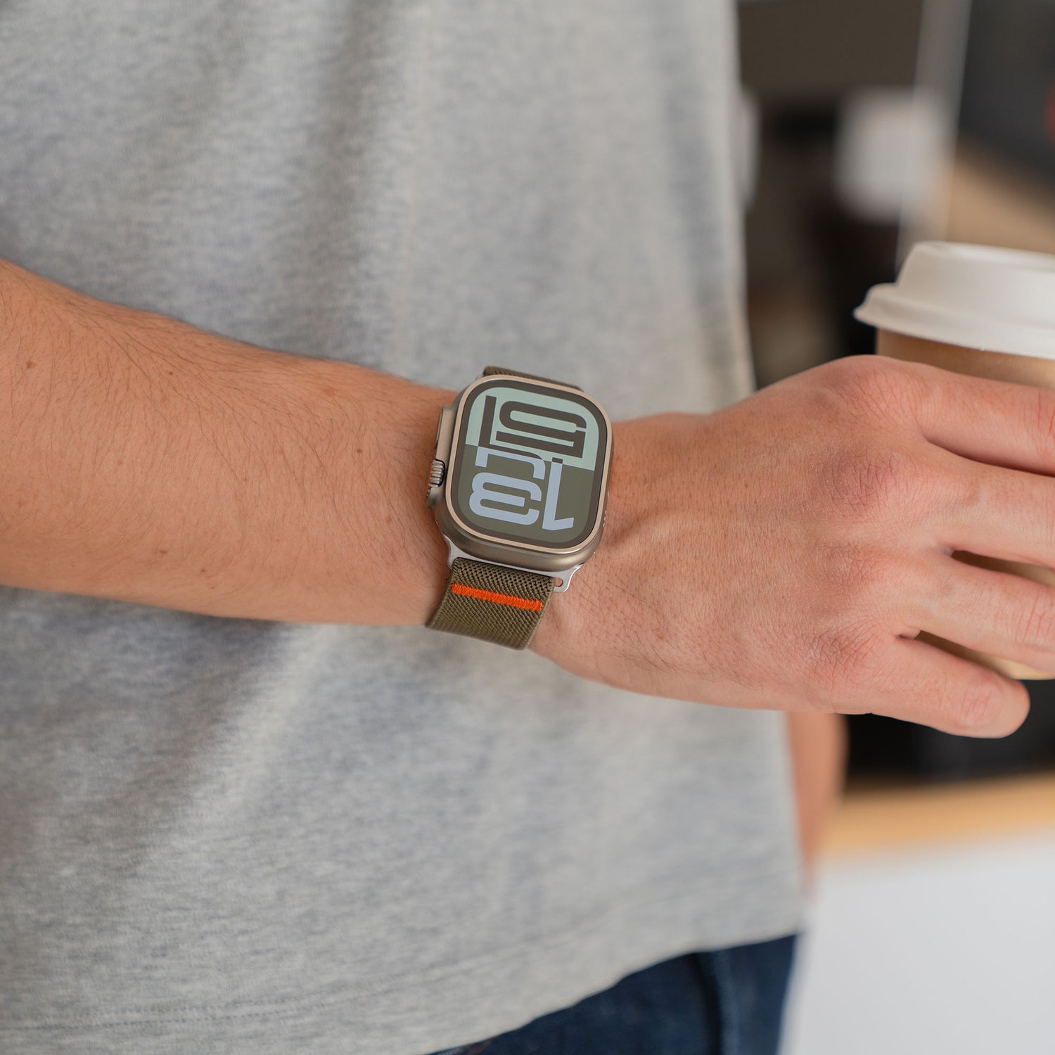 Die Person trägt ein Arktisband Apple Watch Nylonarmband "FlexOne" mit einer großen Digitalanzeige und hält eine Kaffeetasse in der rechten Hand.