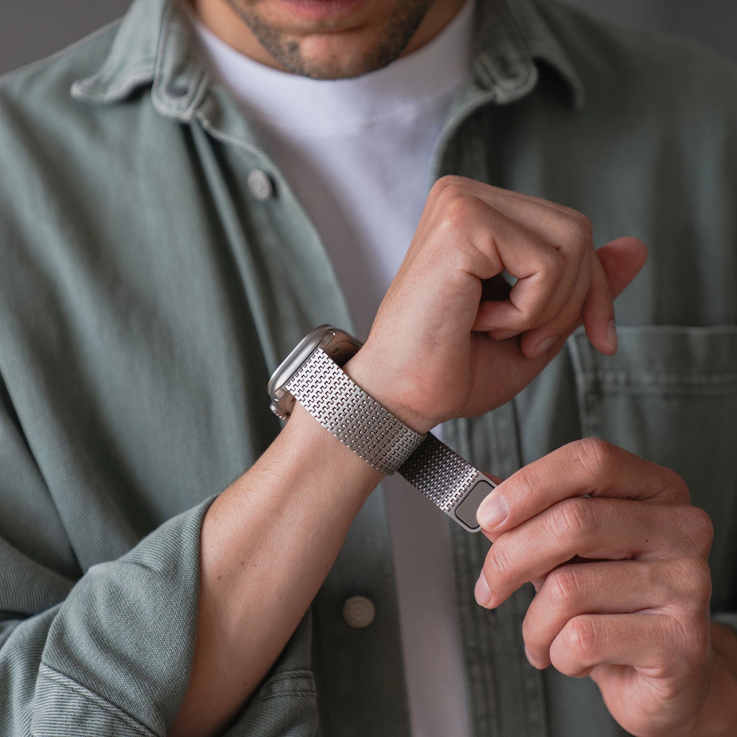 Eine Person in einem hellgrünen Hemd über einem weißen T-Shirt befestigt ein Arktisband Apple Watch Edelstahlarmband „Aurora“ an ihrem linken Handgelenk. Der Fokus liegt auf den Zeigern und der Uhr, wobei das Zifferblatt teilweise sichtbar ist.