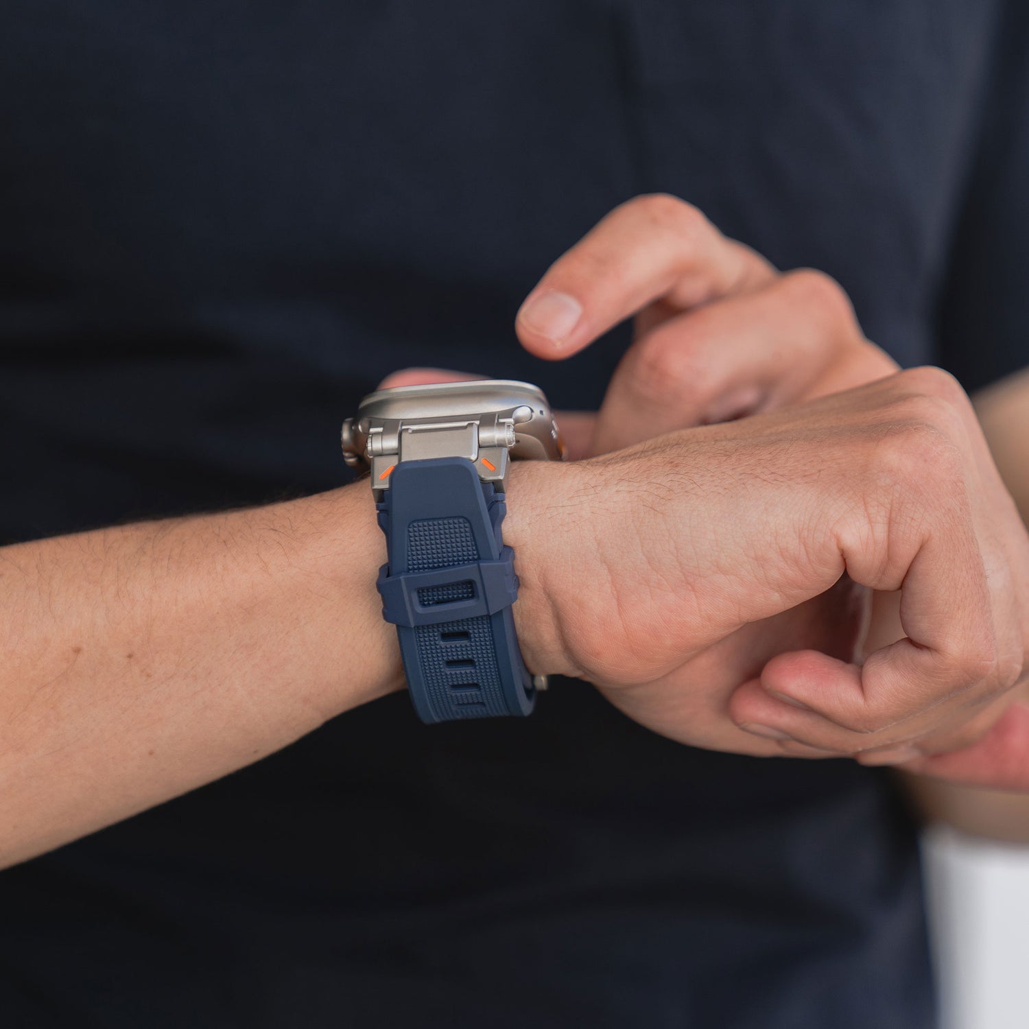 Eine Person in einem dunkelblauen Hemd berührt das Display einer Apple Watch Ultra mit einem Arktisband „Atlas“-Armband von Arktisband am linken Handgelenk. Der schlichte Hintergrund ist unscharf.